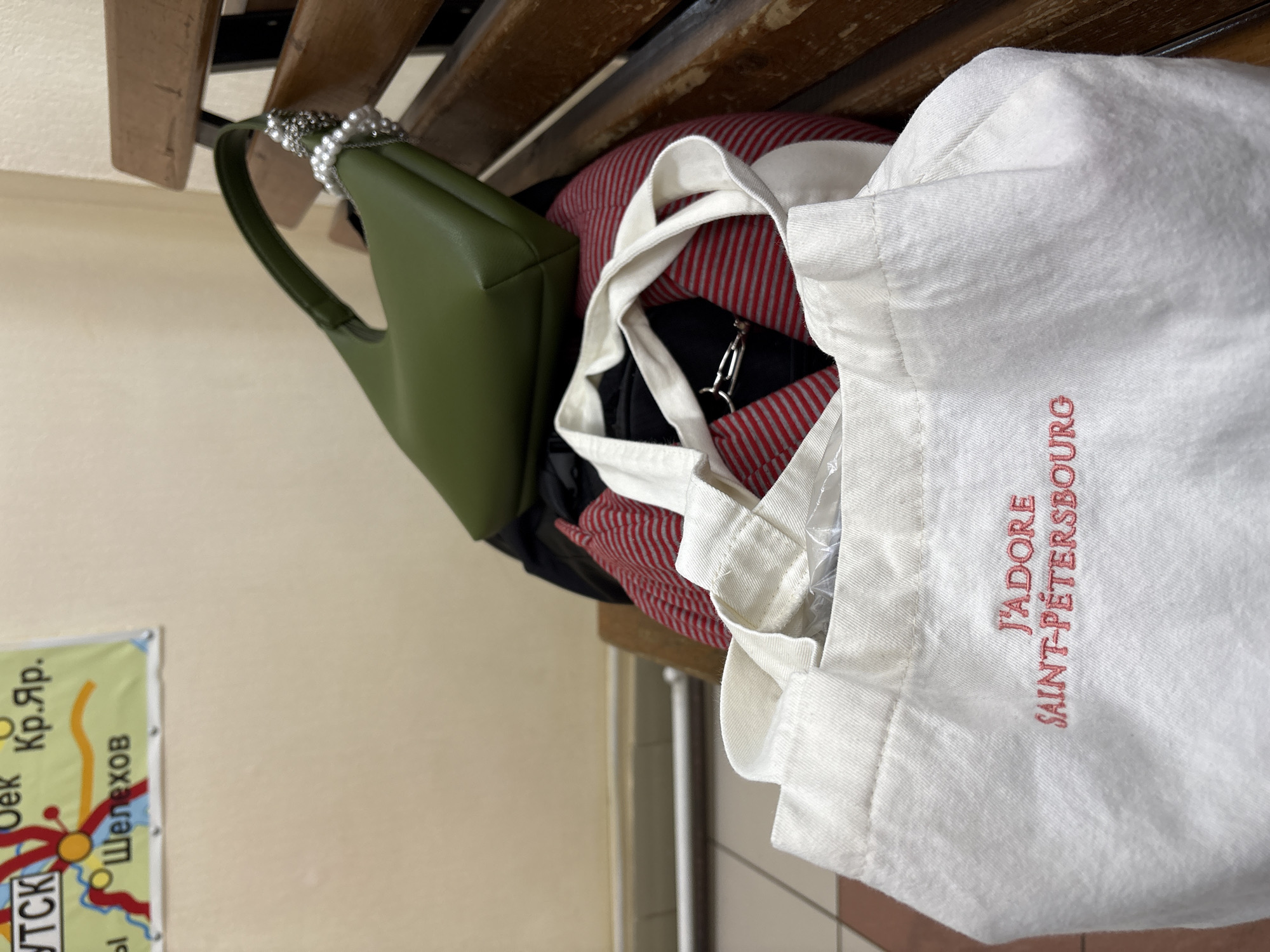 A green purse with a pearl handle, a red and white striped pillow, a black garment, and a white tote bag with the text "J'ADORE SAINT-PÉTERSBOURG" in red embroidery hang from a wooden coat rack. A poster with Cyrillic text and what appears to be a map is partially visible on the left. The location is Ust-Ilimsk, Russia, as indicated by the Cyrillic text. The objects are likely personal belongings placed on the coat rack. The environment is indoors with tiled flooring.