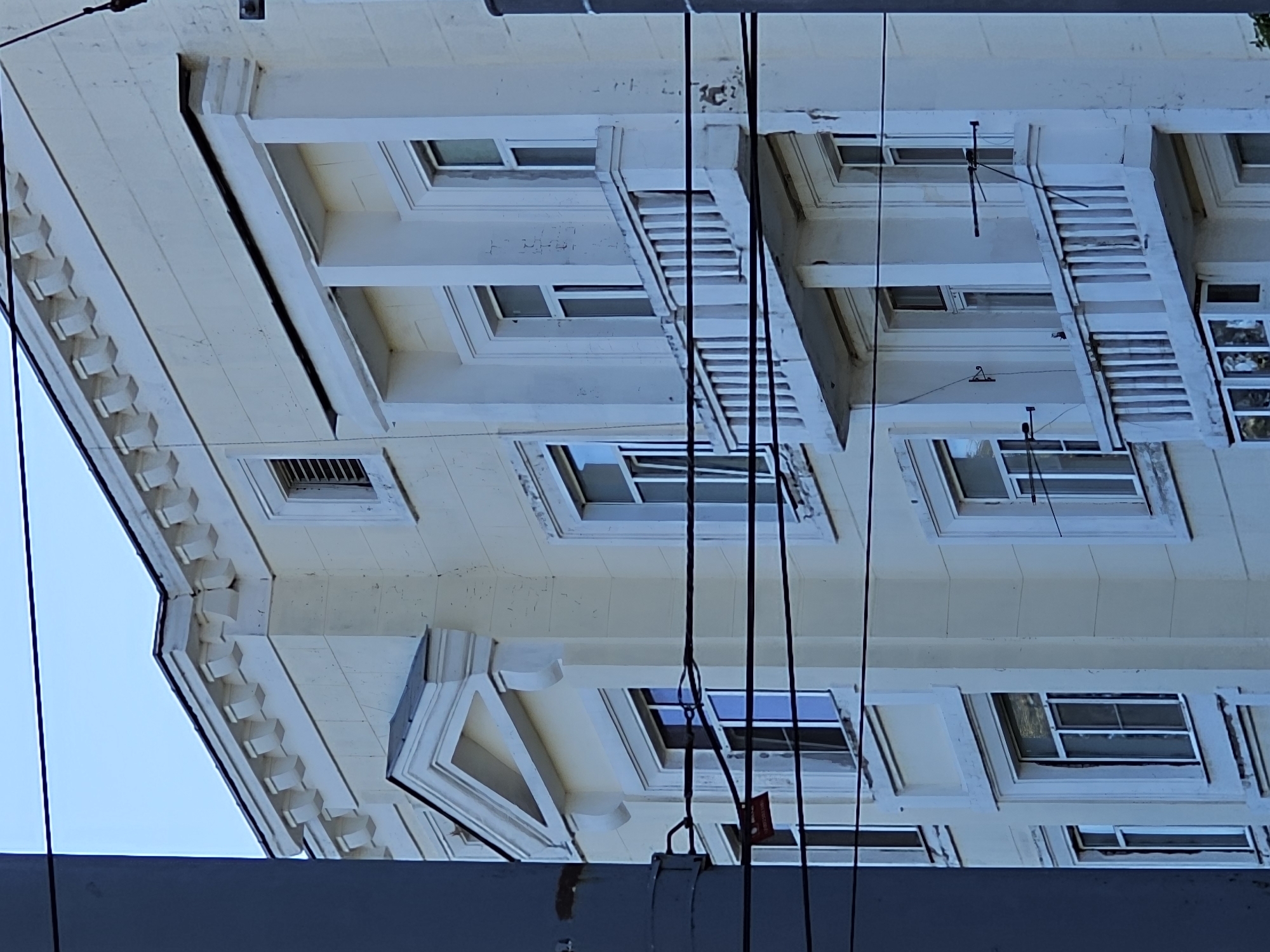This media file captures a low-angle view of a light-colored, multi-story building, likely an apartment complex, seen from below. The architecture features decorative elements such as cornices with repeating geometric patterns and projecting window frames. Several windows are visible, some with their shutters drawn, and a few appear to be slightly ajar. Black electrical or utility wires are prominently strung across the frame, obscuring parts of the building and adding a sense of urban density. The sky is a pale, overcast blue, suggesting it is daytime, possibly in the morning or late afternoon, under diffused lighting conditions. There are no people or discernible human activity in the frame. The scene is static, focusing solely on the architectural details of the building and the overhead network of wires. A small, red tag with white text is partially visible, attached to one of the wires near the bottom center of the image. The overall impression is one of an ordinary urban environment, emphasizing the textured surfaces of the building and the linear elements of the wires against the sky. The orientation of the image is slightly tilted.