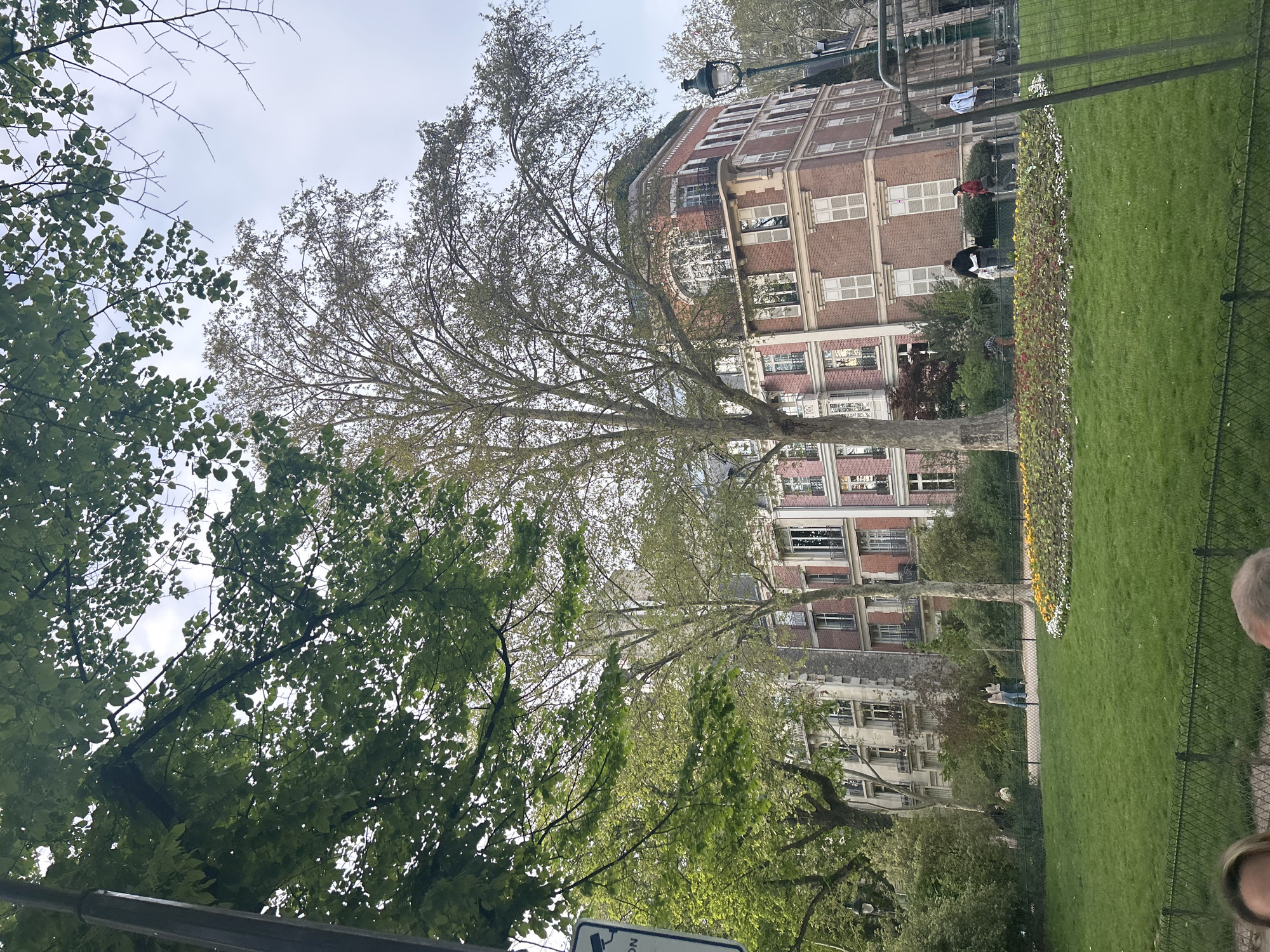 The image shows a view of a Parisian park with a building in the background. The building is a red brick structure with white-framed windows. In the foreground are trees with green foliage and a well-manicured green lawn. A flower bed separates the lawn from a pedestrian pathway where several people are walking. A black metal fence runs along the park's edge. The sky is overcast.