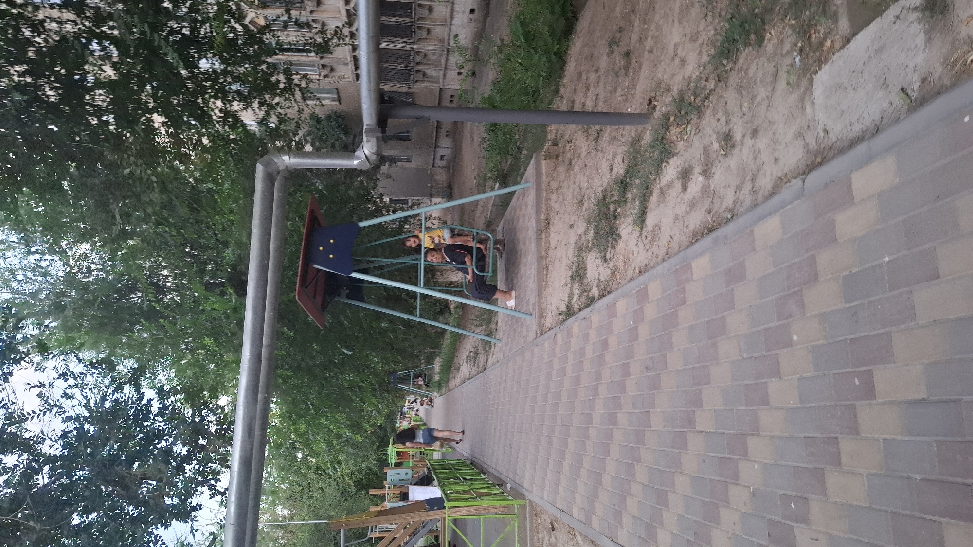 An outdoor shot captures a view of a playground in Shymkent, Kazakhstan. In the foreground, a paved walkway with a brick pattern stretches into the distance. To the right of the walkway, a swing set is visible, with a woman and a young child on it. The woman is seated, wearing dark clothing and white shoes, while the child, in a yellow shirt, is standing on the swing. Behind the swing set, a dirt path and overgrown vegetation are present, leading towards an apartment building. Trees with dense green foliage line the background, casting shadows on the scene. Further down the walkway, another swing set and playground equipment are visible, suggesting a larger recreational area. A woman in shorts and a dark top is walking away from the camera on the walkway. The overall impression is of a quiet, residential area with a focus on outdoor recreation.