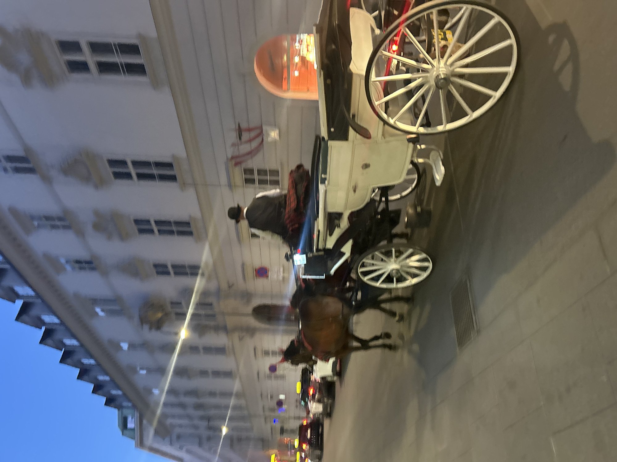 The image shows a horse-drawn carriage in Vienna, Austria, at dusk. The carriage is white with a dark interior and has large, spoked wheels. A person wearing a hat and a vest is seated in the driver's position, and a horse is pulling the carriage. The carriage is on a cobblestone street, and several cars are visible in the background. The buildings lining the street are typical of Viennese architecture, with ornate facades and windows. The sky is a deep blue, indicating the early evening. The overall scene evokes a sense of old-world charm mixed with modern city life.
