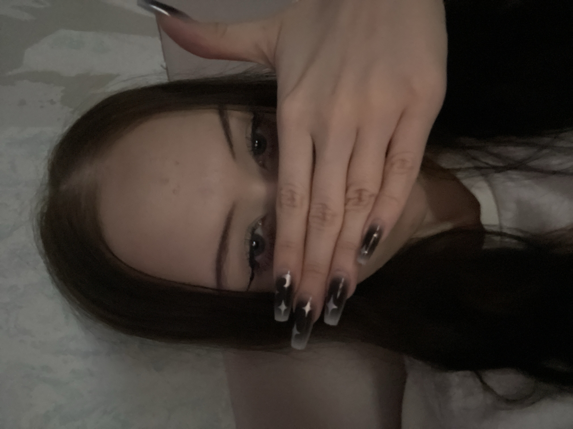 The media file shows a close-up, angled shot of a person's face and hand, with their hand partially obscuring their face. The person has dark hair and is wearing makeup, with one eye visible. Their fingernails are long and painted with a dark, gradient design featuring stars and a crescent moon. The background appears to be a light-colored, textured surface, possibly a bed or sofa. The lighting is dim, suggesting an indoor setting, possibly at night or in a low-light environment. The overall mood is somewhat mysterious and perhaps a bit edgy due to the makeup and nail art. There is no visible text in the image. The location context is Votkinsk, Russia, but there are no specific visual cues in the image to confirm this.