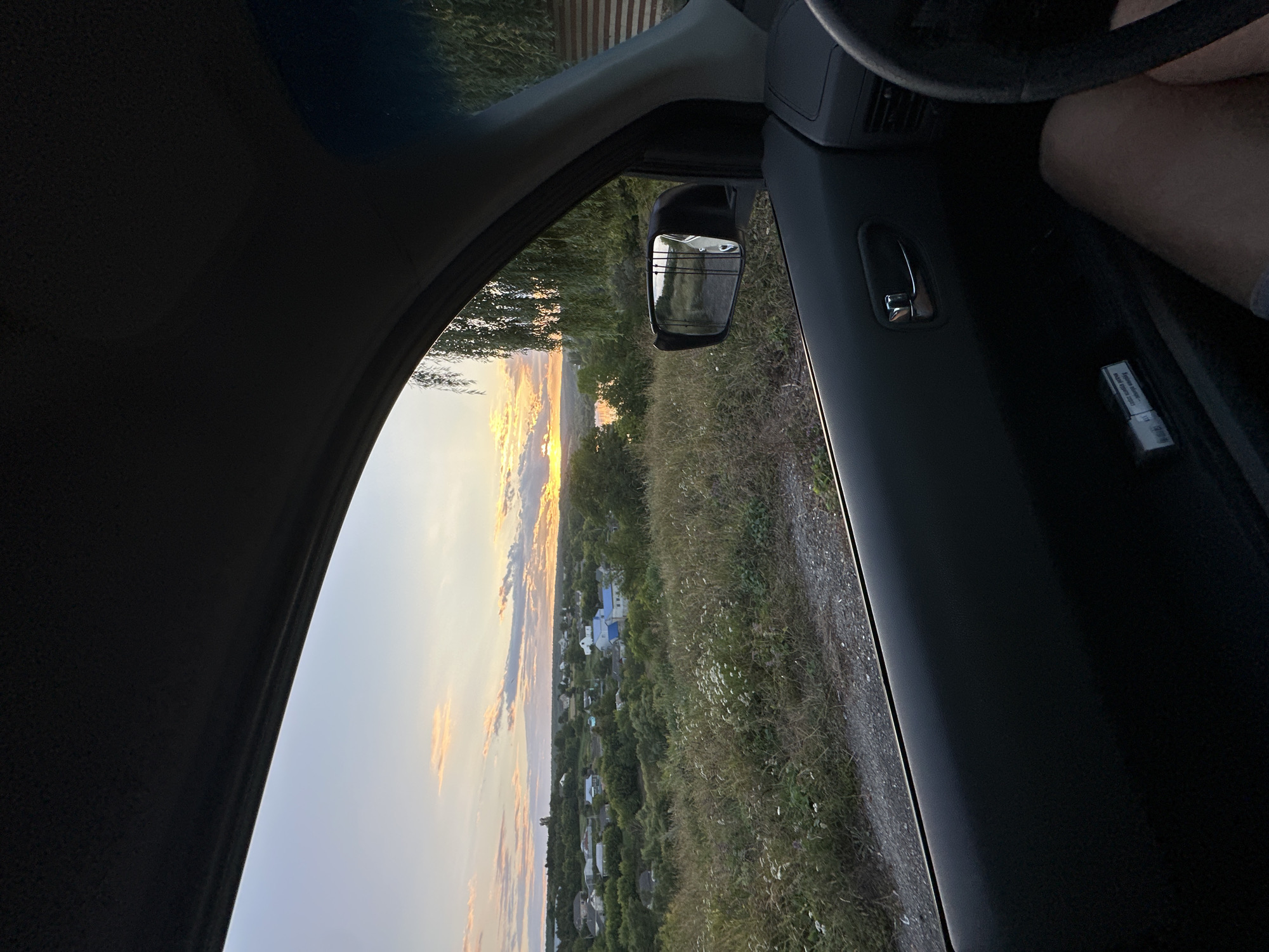 The image, taken from inside a vehicle, offers a view of a sunset over a rural landscape near the city of Makiv, Ukraine. The perspective is from the passenger seat, looking out the open window. In the foreground, the car's interior is visible, including the door panel with a chrome handle and a pack of cigarettes on the lower ledge. A person's leg, clad in shorts, is visible on the right side of the frame.
The scene outside the window depicts a village nestled amongst rolling hills and dense greenery. The sky is painted with hues of orange, pink, and yellow as the sun dips towards the horizon, casting a warm glow over the landscape. A body of water, possibly a river or lake, is visible in the distance. Tall trees, likely willows, with their characteristic drooping branches, frame the right side of the view. The ground in the foreground appears to be a dirt or gravel road. The overall impression is one of a peaceful, rural evening.