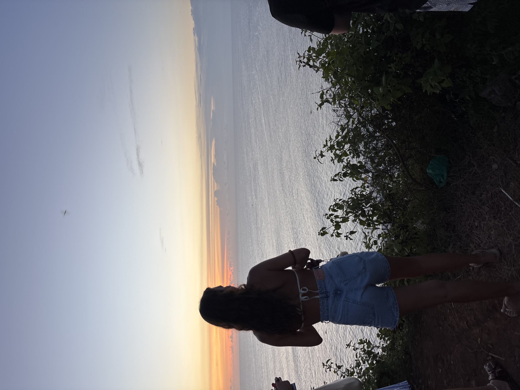 Captured in Pecatu, Indonesia, the image presents a twilight scene where a woman stands silhouetted against a horizon transitioning from a vivid orange sunset to a muted blue sky above a body of water, presumably the ocean. The woman, clad in denim shorts and a light-colored top, is positioned near the edge of vegetation, with glimpses of plant life on her right. In the distance, a small aircraft is visible in the sky. The overall composition suggests a viewpoint from a cliff or elevated location.