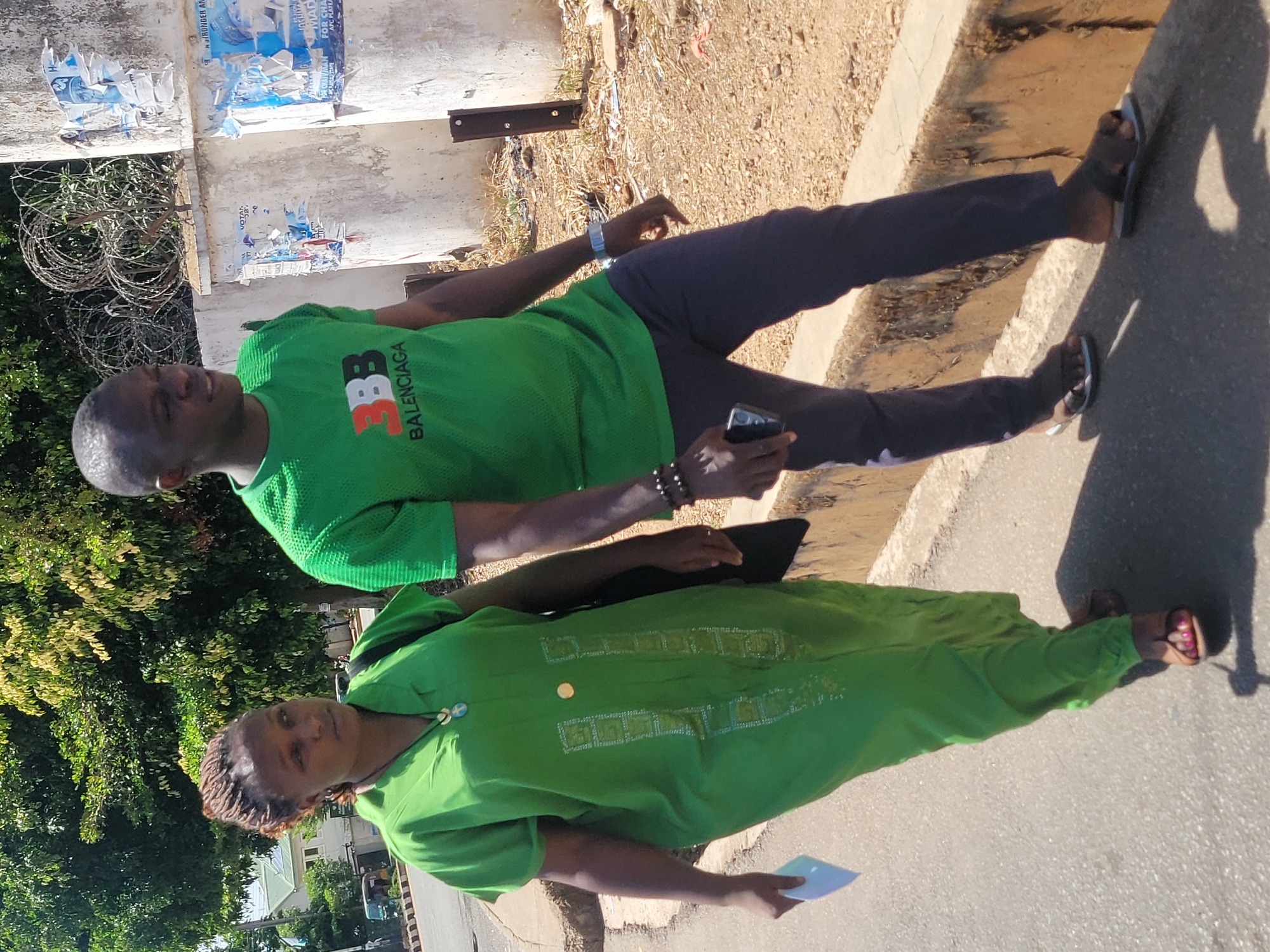The image shows two individuals in Jos, Nigeria. A man in a green "Balenciaga" t-shirt and dark pants is standing next to a woman wearing a long green dress. The man is holding a cell phone. The woman is holding a white card. They are standing on a paved area next to a curb. Behind the man is a building with posters and barbed wire. A tree is on the left.