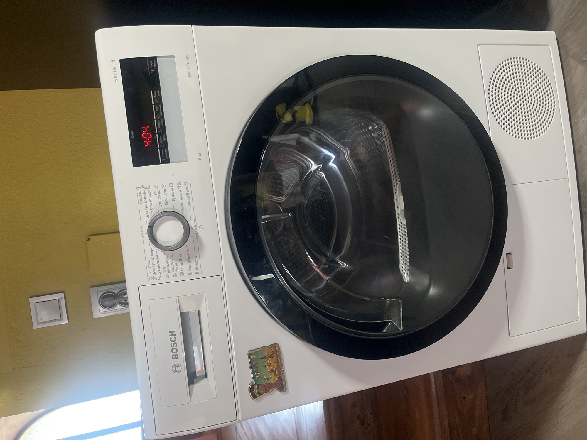 A Bosch Series 4 Heat Pump dryer is shown in a room. The dryer is white and has a large glass door. The control panel has a digital display showing "4:04", and various program selection buttons. The dryer is labeled "8 kg". A decorative magnet depicting ancient Egypt is attached to the left side of the dryer. The background of the image shows a yellow-painted wall with a light switch and an electrical outlet. The floor is wooden. This scene is located in the city of Tarnivtsi, Ukraine.