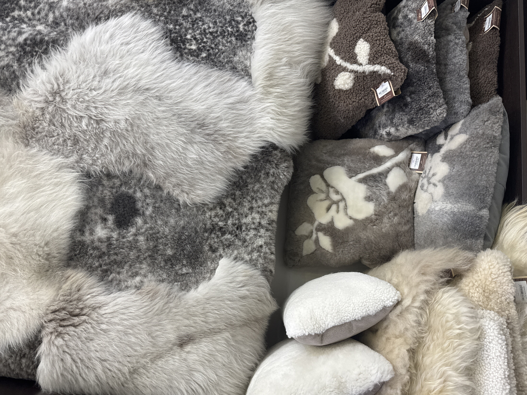 The image shows a close-up, overhead view of a collection of plush, fluffy sheepskin rugs and pillows. The items are predominantly in shades of gray, white, and beige, with some featuring floral patterns in contrasting colors. They are piled on top of each other, creating a textured and luxurious display. The scene appears to be in a retail setting, likely a shop or market stall, given the presence of price tags on some of the items. The overall impression is one of comfort and softness. There are no people visible in the image. The location is not specified in the image itself.
