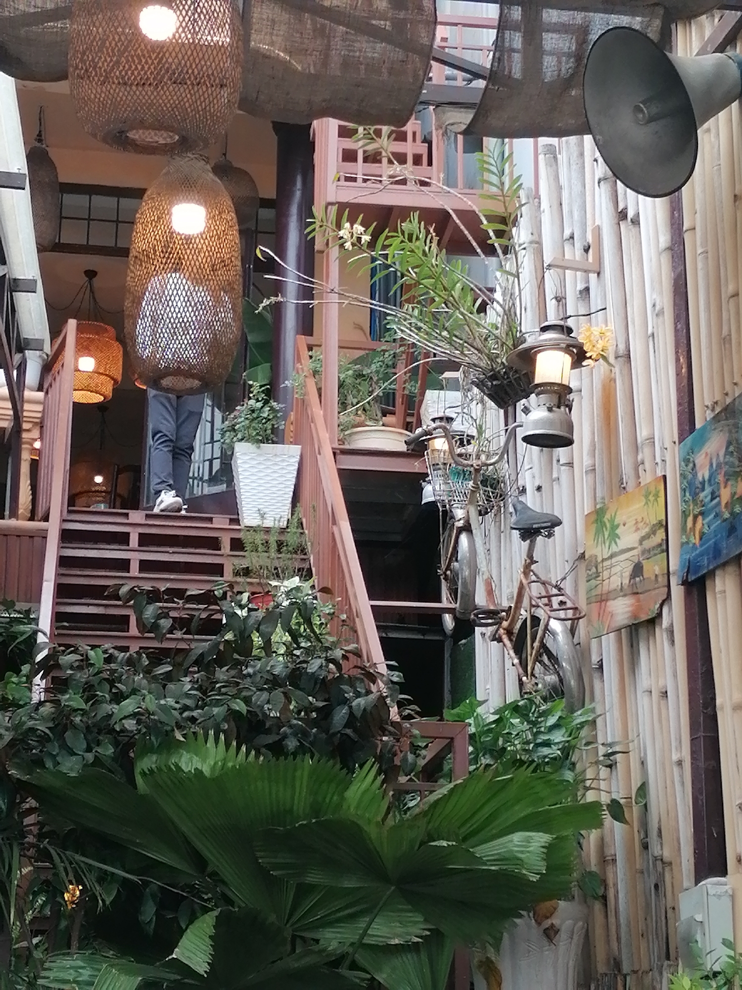 The image captures an outdoor cafe or restaurant setting in Da Lat, Vietnam. The scene is dominated by lush greenery and natural materials. Large, woven pendant lights hang from the ceiling, casting a warm glow. A person is standing on a wooden staircase, wearing dark pants and light-colored sneakers, appearing to be ascending or descending. To the right, a wall constructed of vertical bamboo stalks serves as a backdrop. A vintage bicycle, adorned with a lantern and a basket containing plants, is leaned against the bamboo wall. Decorative paintings featuring natural landscapes are mounted on the wall. The overall ambiance suggests a rustic and tropical atmosphere, characteristic of many establishments in Da Lat.