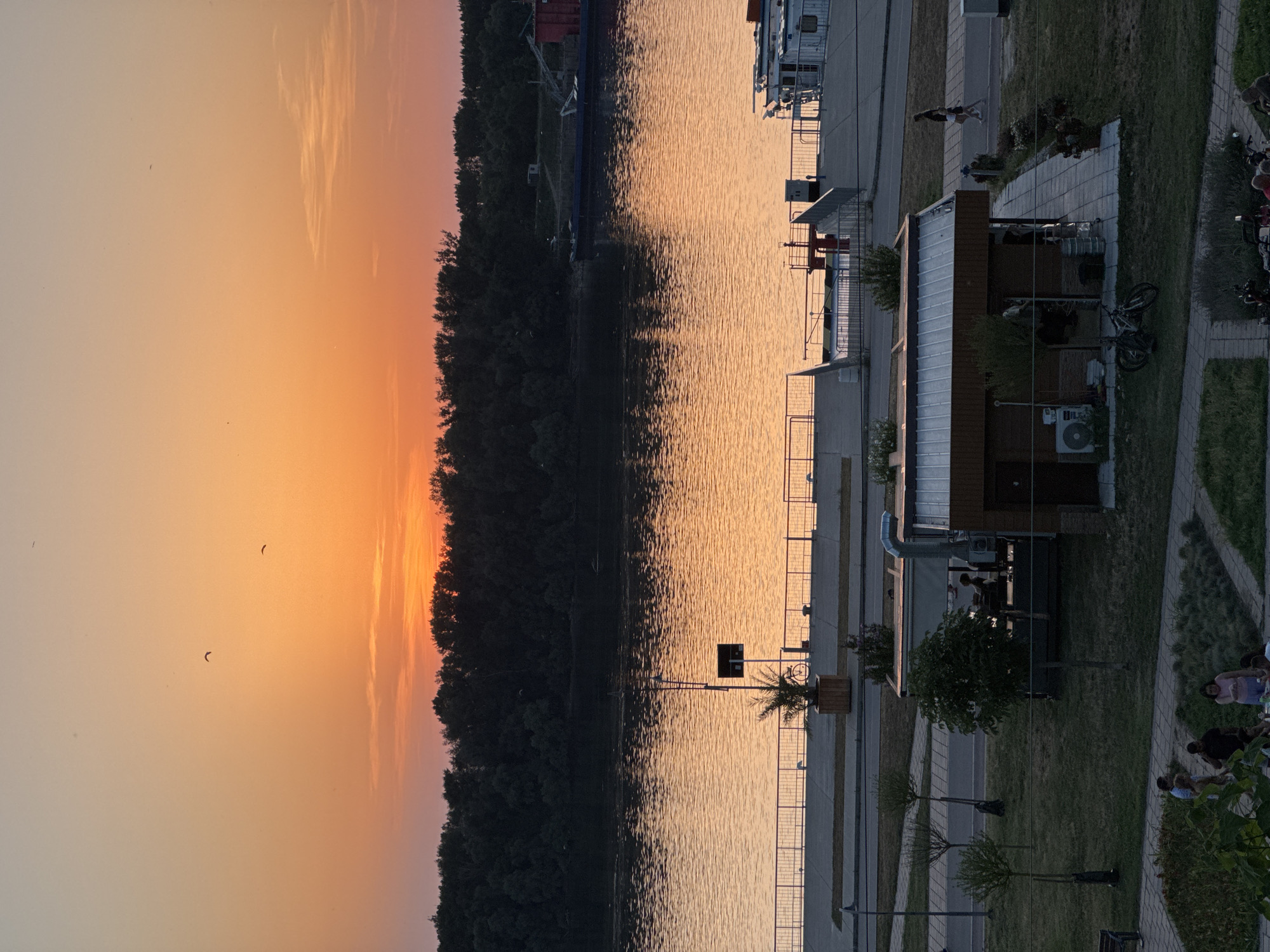 The image depicts a sunset over the Danube River in Ruse, Bulgaria. The sky is a gradient of orange and yellow, with a silhouetted treeline across the river. A wide, calm river reflects the sunset's colors. Along the riverbank in the foreground, there is a walkway with a metal railing. Several buildings, including a small kiosk-like structure with outdoor seating and bicycles parked nearby, are visible on the grassy area behind the walkway. People are seen walking and sitting in small groups in the foreground and on the walkway. In the distance, on the river, a large barge is docked. The overall scene is peaceful, with the warm hues of sunset creating a serene atmosphere.