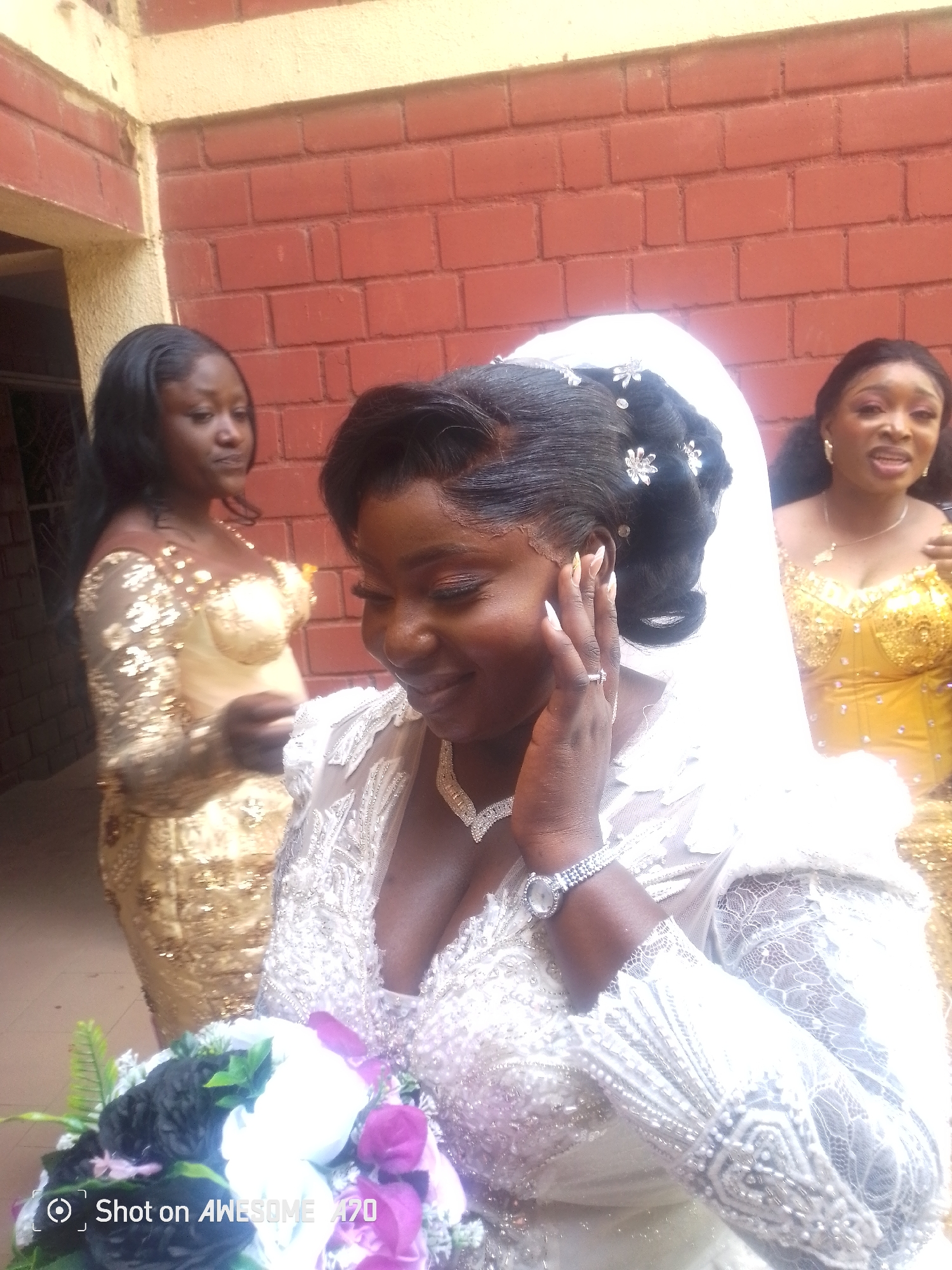The image depicts a wedding scene, likely in Kuru Karama, Nigeria. The main subject is a bride in a white, heavily embellished gown and veil. She is holding a bouquet of flowers in various colors. She is wearing a watch and a diamond necklace, and has her hand near her face, possibly touching her ear. Two other women are visible in the background. One is wearing a gold, sequined dress and the other, a yellow sequined dress. They appear to be bridesmaids or guests. The background suggests an outdoor setting, possibly near a brick building. The image has been taken with a phone, as indicated by the watermark "Shot on Awesome A70".
