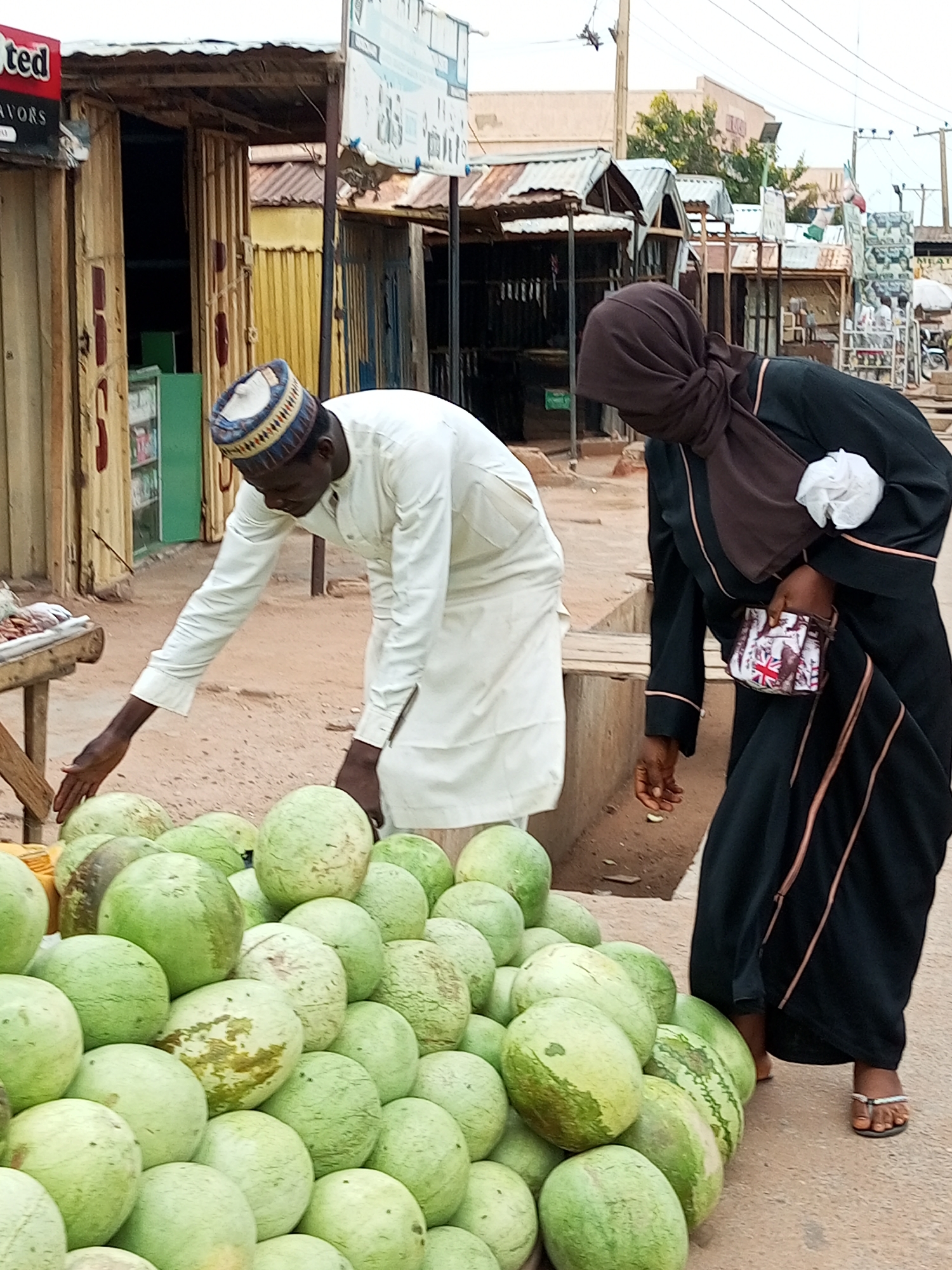 A man wearing a white robe and a patterned cap is reaching towards a pile of watermelons at a market stall in Gombe, Nigeria. A woman wearing a dark hijab and a black abaya is standing behind him, also looking at the watermelons. She is holding a small purse with a Union Jack pattern. The market appears to be outdoors, with stalls made of corrugated metal and wood. The ground is dusty, and other market stalls and buildings are visible in the background.
