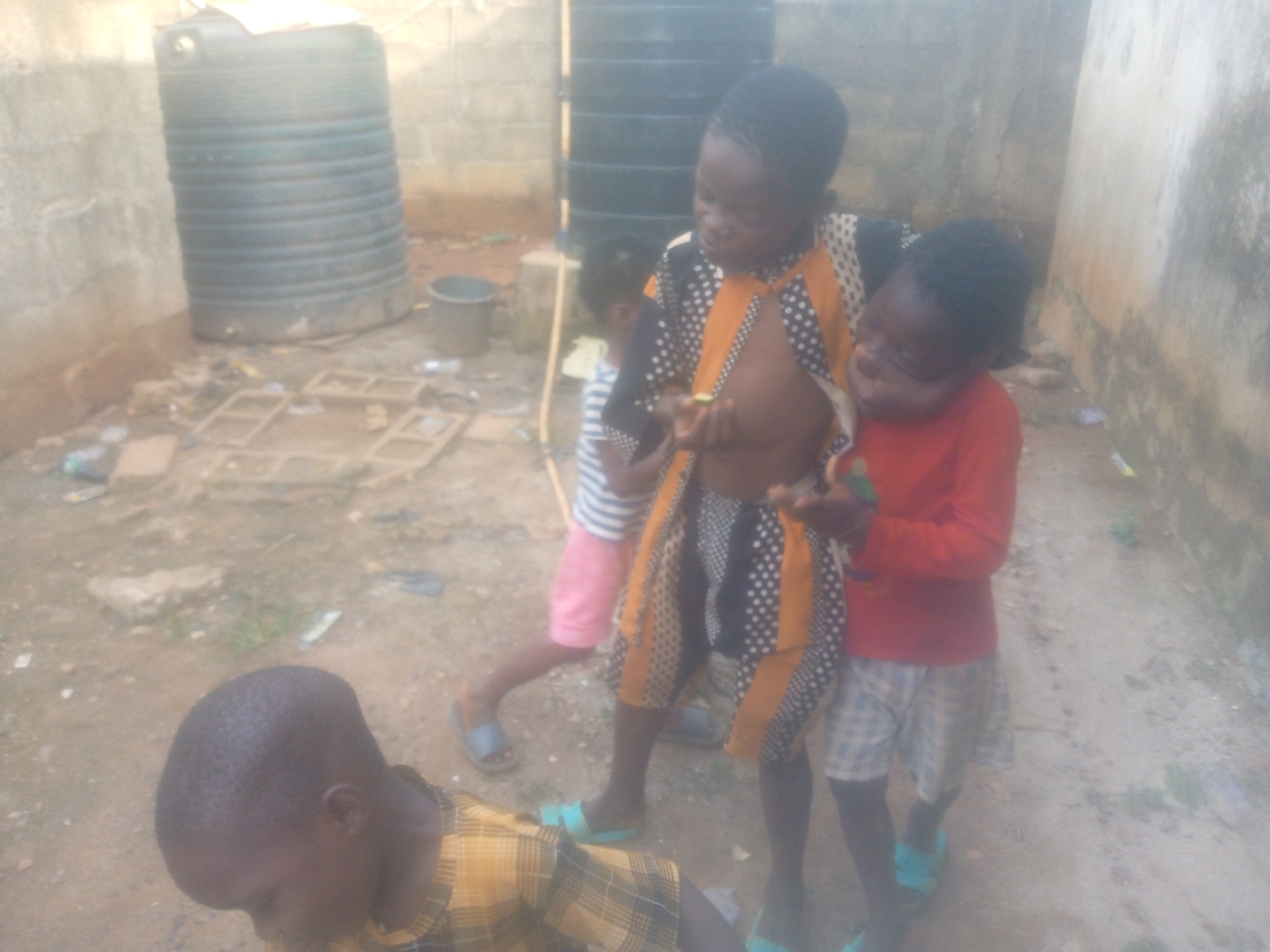 Four children are gathered in what appears to be a dusty, outdoor area with concrete block walls in the background. The ground is dirt, littered with scattered debris including broken bricks and what looks like trash. A large, black cylindrical water tank stands prominently in the background, near a smaller black bucket.
In the center of the image, one child wears an open, black and orange patterned shirt, revealing a bare torso, and light blue flip-flops. This child is holding a small, indistinct green object in their hands and is looking down at it, with a slight smile. To the right of this child, another child wears a red long-sleeved shirt and plaid shorts, also with light blue flip-flops, looking towards the central child. Partially visible to the left and slightly behind the central child is a third child, wearing a striped top and pink shorts with dark flip-flops. In the lower foreground, a fourth child is partially visible, wearing a yellow and black plaid shirt and looking downwards.
The scene is lit by natural daylight, but the image has a soft, slightly hazy quality, which could be due to atmospheric conditions or camera focus. The children appear to be interacting casually, possibly playing or simply observing the object held by the central child. There is no visible text in the image.