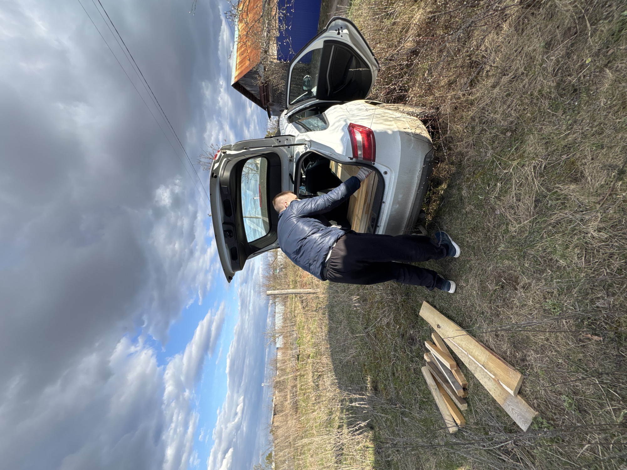 A man is depicted in a rural or semi-rural outdoor environment in Noviy, Russia, actively engaged with the open trunk of a white car. He is leaning into the car's rear, appearing to load or arrange rough-cut wooden planks, several of which are already visible inside the trunk. Additional wooden planks lie scattered on the dry, sparse grassy ground beside the vehicle.
The car, a white hatchback or small SUV, shows signs of dirt or mud on its lower body, suggesting travel on unpaved surfaces. The man is dressed in a dark blue puffer jacket, dark trousers, and athletic shoes, and wears light-colored gloves on his hands.
The setting features an expanse of dry, light-brown grass and leafless bushes, indicating either early spring before new growth or late autumn. A rustic building with a rusty corrugated metal roof and blue siding is visible in the background. The sky is partly cloudy, with significant patches of blue sky peeking through dark grey clouds. Overhead, power lines traverse the scene. The overall impression is of a functional activity taking place in a utilitarian, outdoor area during daytime. No discernible text is visible.