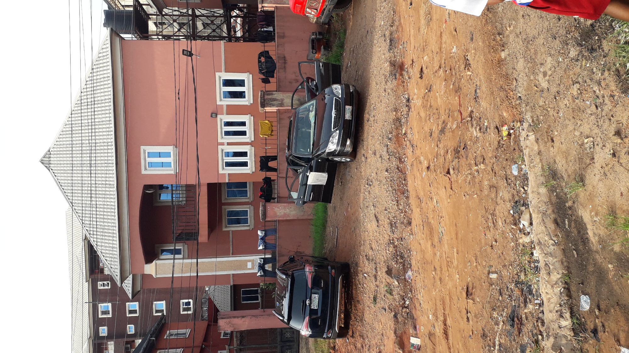 The image depicts a scene in Nkpor, Nigeria. A three-story salmon-colored building with white-framed windows is visible. Clothes are hanging on the balconies. Two black cars are parked in front of the building, one with an open door. A red vehicle is partially visible near the building. The ground is a mix of reddish-brown dirt, gravel, and trash. Power lines run in front of the building. Part of a person wearing a red shirt and a white shirt is visible in the top right corner.