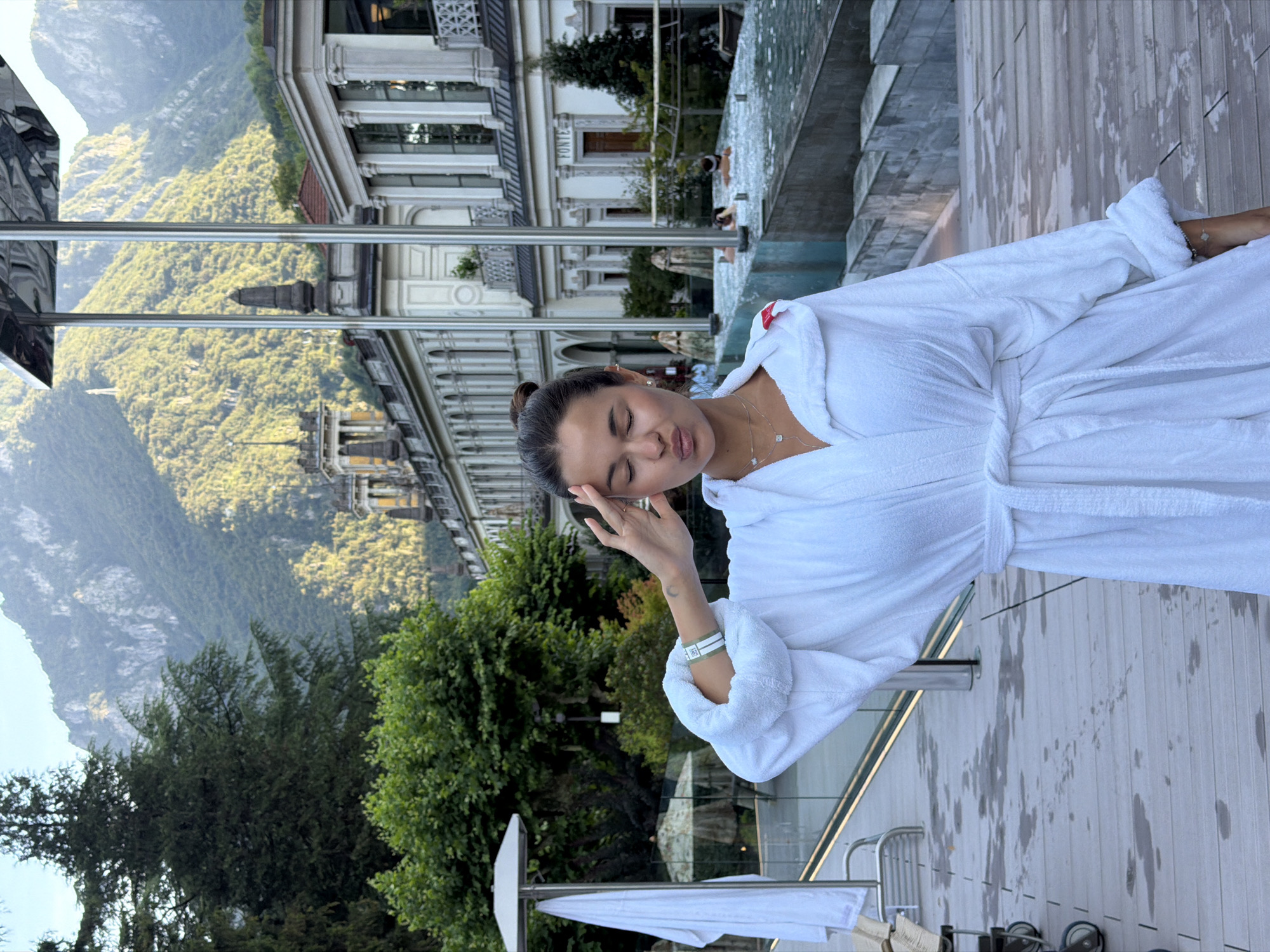 In the city of San Pellegrino Terme, Italy, a young woman stands on a wooden deck beside an outdoor pool. She is wearing a white bathrobe and has her eyes closed, with her hand touching her forehead. The background features lush green mountains, a historic building with classical architecture, and several people relaxing in the water. The scene appears to be a serene spa environment.