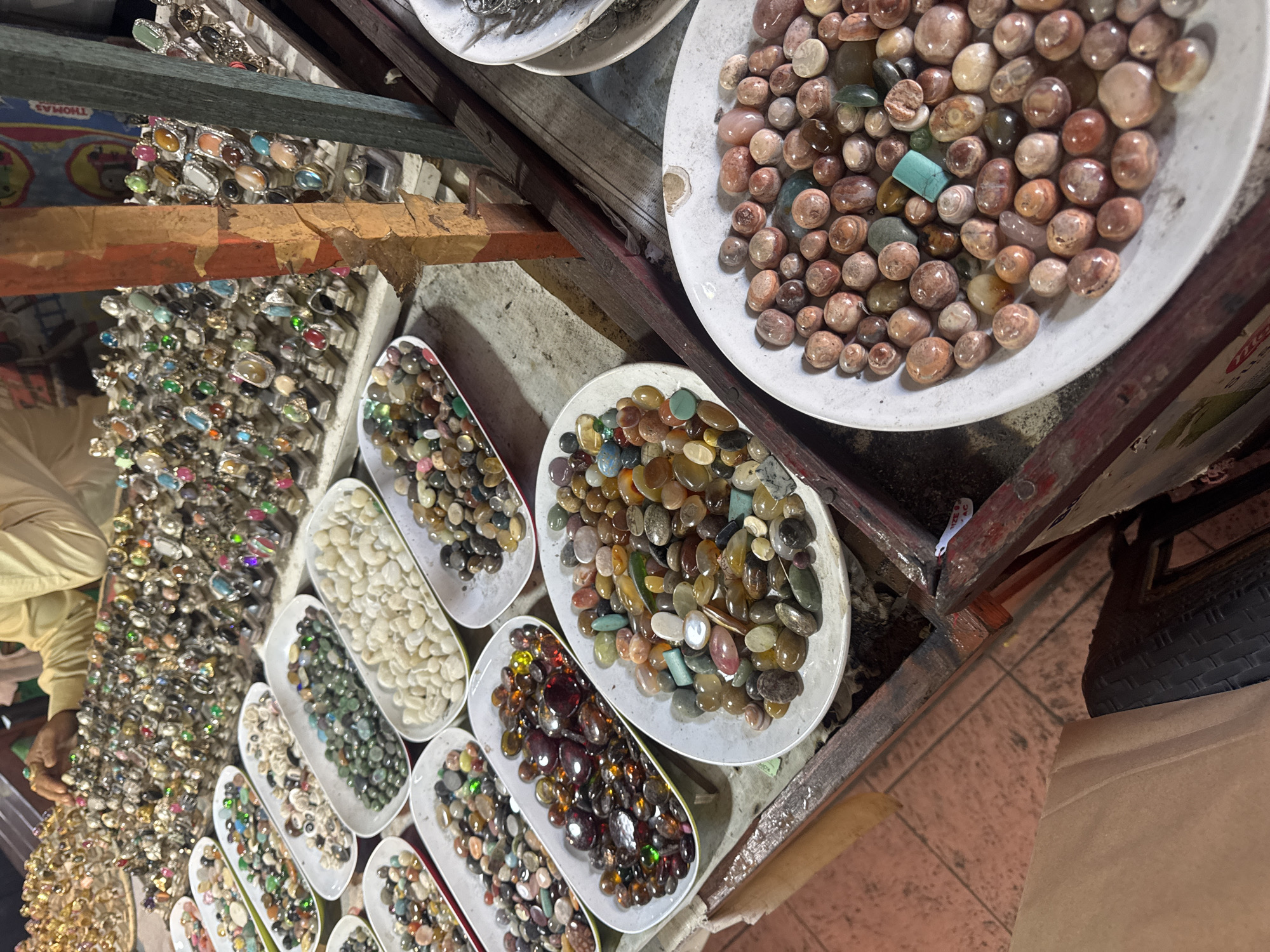 A street market stall in Denpasar, Indonesia, displays an array of colorful stones and rings. The foreground features several white oval trays filled with polished, tumbled stones in various shades of brown, red, green, white, and blue. To the left and slightly behind these trays, a tray of dark red and brown stones is visible, and further back, trays of white and green stones are arranged. The background shows rows of rings on display stands. A person wearing a light-colored garment is partially visible, reaching towards the rings. The stall has a rustic wooden structure with red and brown painted posts. The ground appears to be tiled.