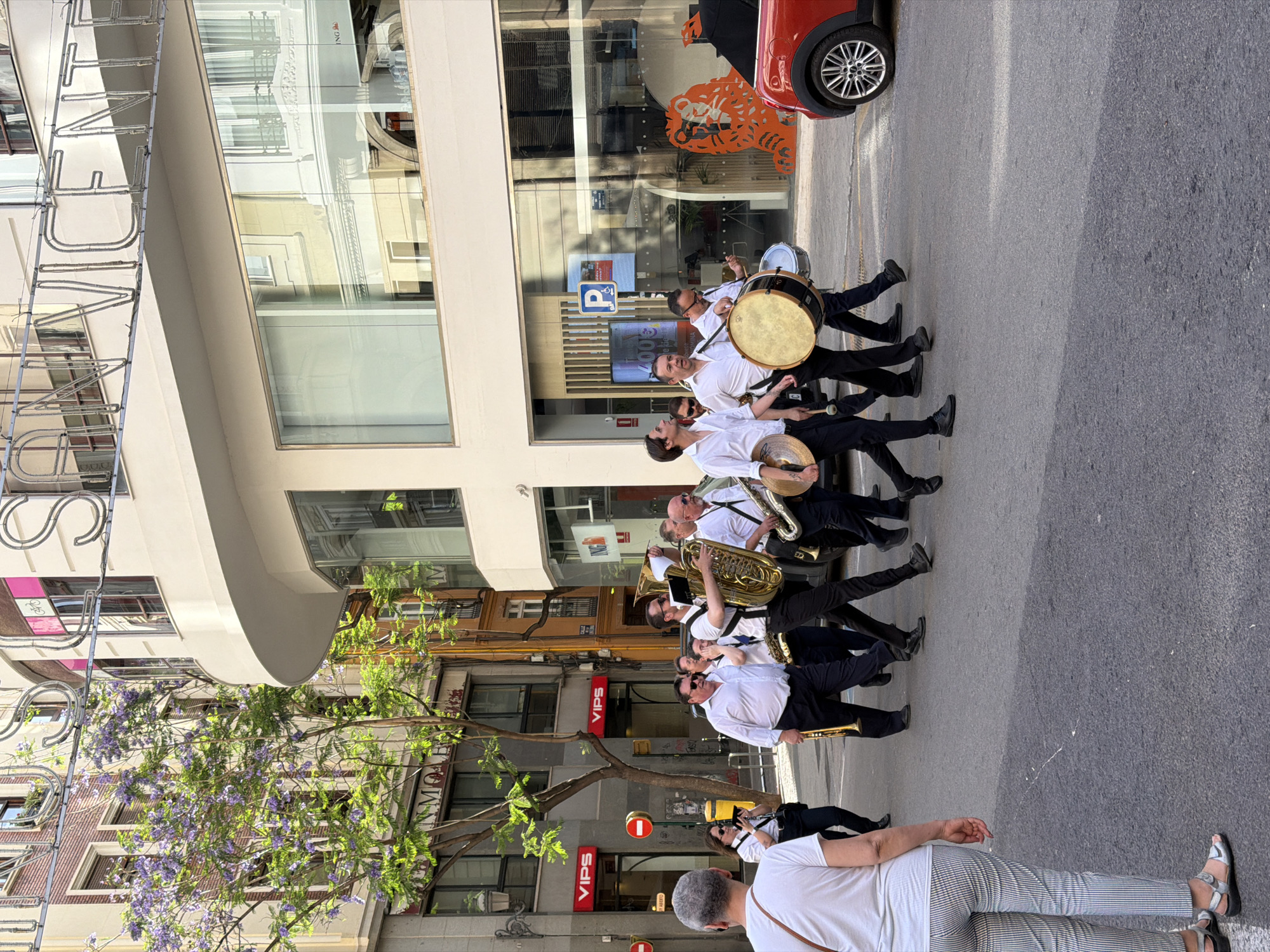 A street band is marching in a city street. The band members are wearing white shirts and dark pants. They are playing various instruments, including a tuba, a saxophone, a trumpet, and drums. The band is walking on the right side of the street, towards the viewer. In the background, there are buildings with shops on the ground floor. One of the shops has a sign that says "VIPS". There is also a red car parked on the side of the street. The street is paved with asphalt and there are some trees and streetlights. The sky is blue and there are some clouds. The image was taken in València, Spain, as indicated by the neon sign in the background that reads "SAN VICENTE". The overall scene suggests a festive event or parade.