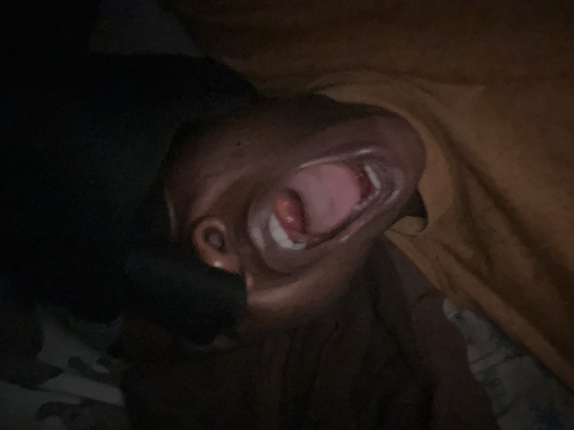 The image is a close-up, low-angle shot of a person's face with their mouth wide open, as if yawning or screaming. The lighting is dim, with the face illuminated from the front, creating shadows that obscure details. The person's eyes are visible, and their tongue can be seen inside their mouth. They appear to be wearing a light-colored t-shirt. The background is dark and out of focus, making it difficult to discern any specific environmental details. No specific location in Ikorodu, Nigeria can be identified from this image.