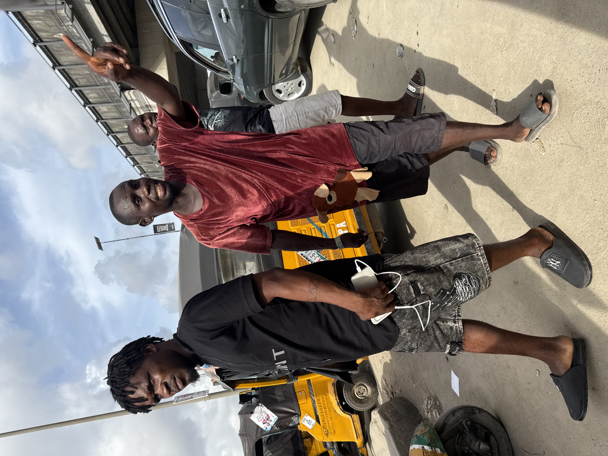 Three Black men stand posing in front of an auto rickshaw in Lekki, Nigeria. One man in the foreground wears a black T-shirt and patterned shorts. He holds a white charger. The second man wears a maroon shirt and black shorts and is smiling. The third man is behind the second and is pointing upwards. The auto rickshaw is yellow and has writing on it. A car is parked behind them. Above them is an elevated road. The sky is cloudy.