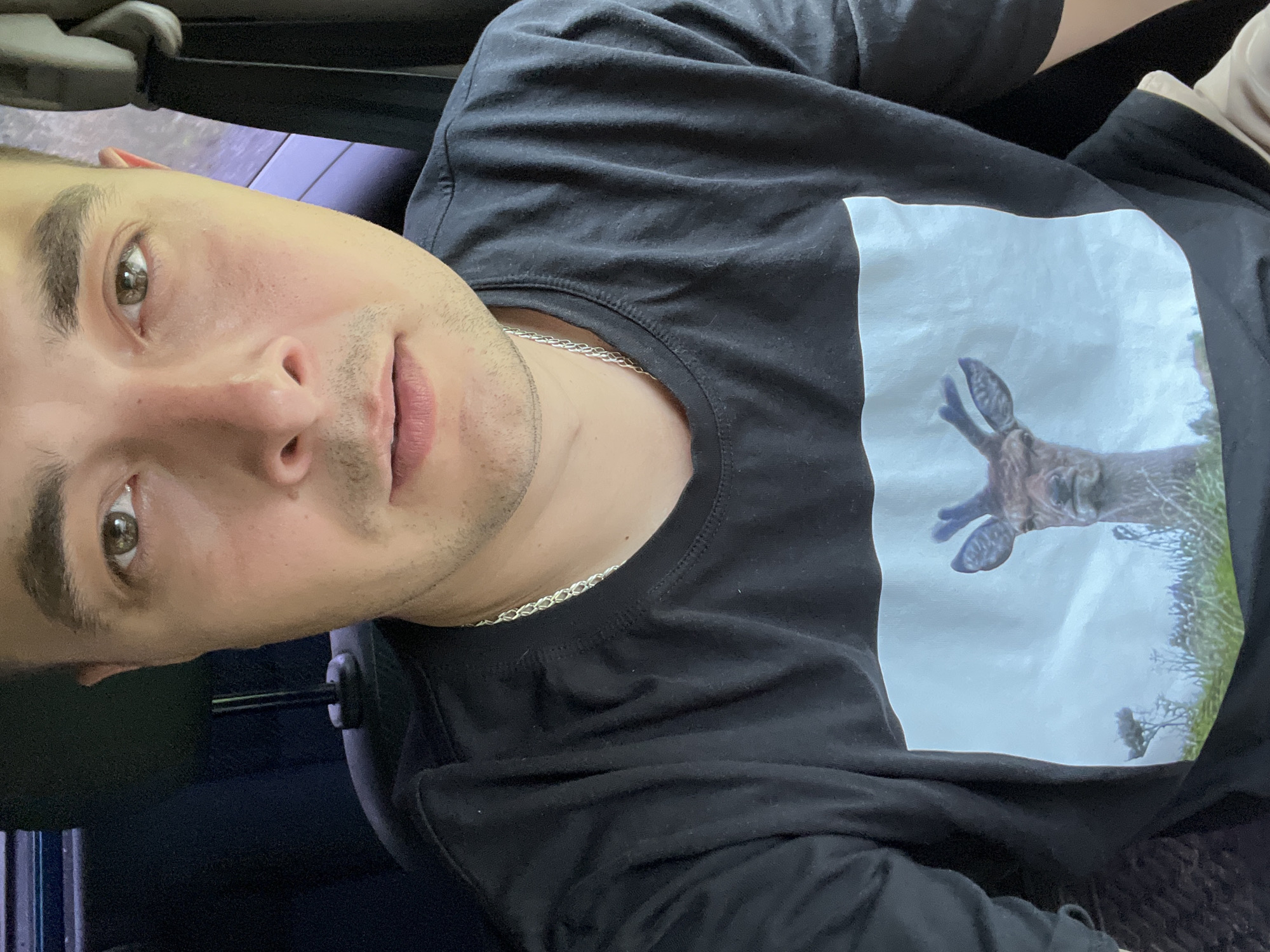 A man, appearing to be of Caucasian descent, is positioned inside what appears to be a vehicle. He is looking directly at the camera, his expression neutral. He is wearing a black t-shirt with a printed image of a deer's head. A silver chain necklace is visible around his neck. The background is dark and blurred, suggesting the interior of a car with some ambient light. The provided context indicates the location is Razdolye, Russia.