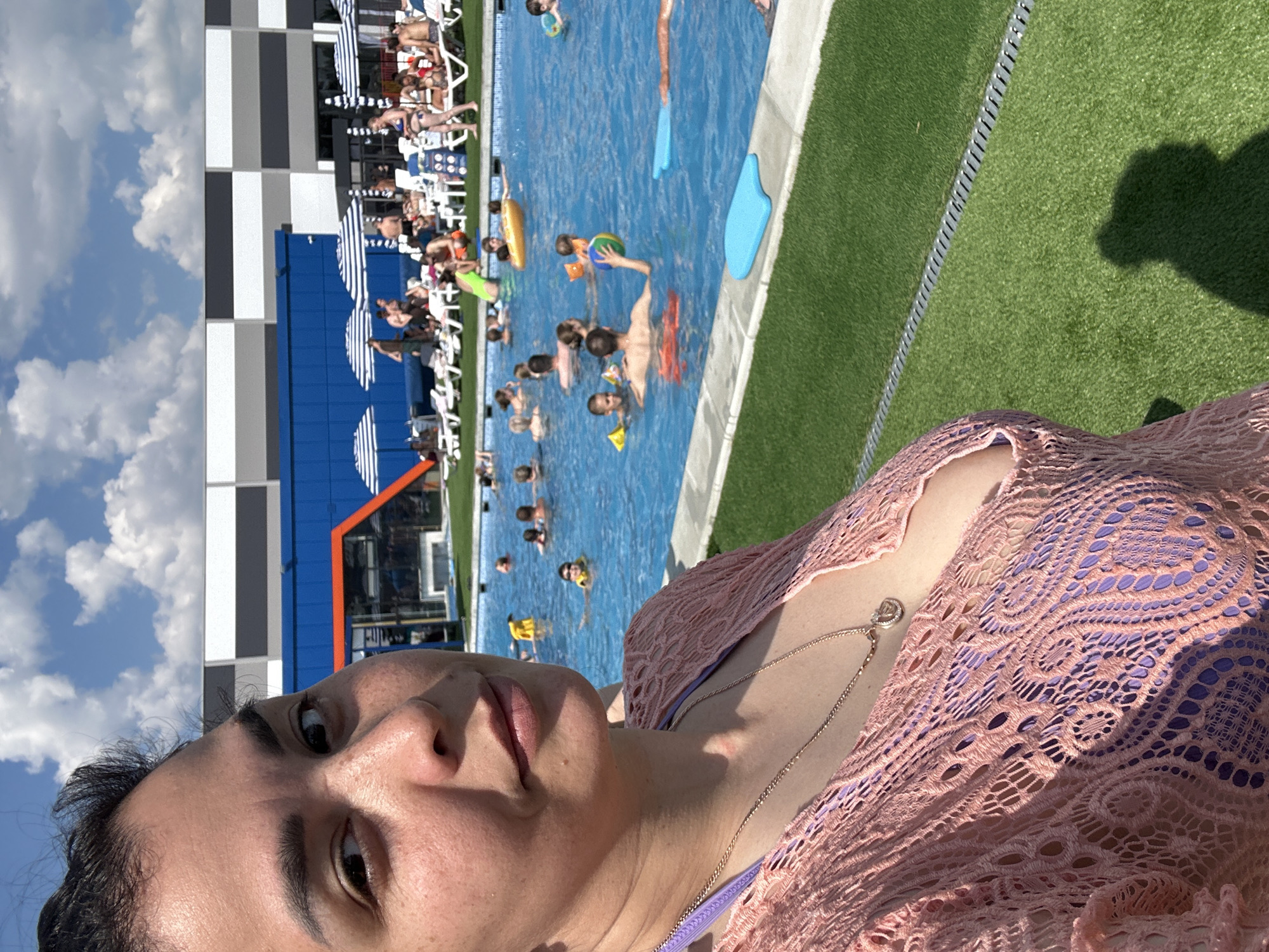 In an outdoor swimming pool area, a woman in the foreground takes a selfie. She is wearing a pink lacy cover-up over a purple swimsuit and a necklace with a heart-shaped pendant. Behind her, the blue water of the pool is filled with people of various ages swimming and relaxing on inflatable toys. Some are in the water, while others are near the edge, interacting with one another. Deck chairs and striped umbrellas are visible along the poolside, and a modern building with blue and grey sections forms the backdrop. The ground surrounding the pool is covered in green artificial turf. The sky above is clear with scattered white clouds.