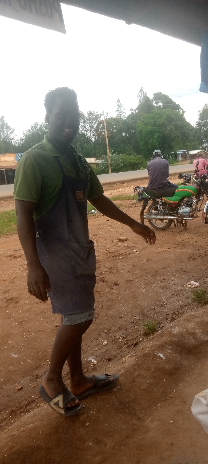 A young Black man wearing a green polo shirt, a blue apron, and black sandals is smiling and standing on a dirt path in front of a small store in Kivaywa, Kenya, near a motorcycle parked along the roadside. He is facing the camera. Behind him are trees. To the man's right, a Black man wearing a grey shirt and a black cap is sitting on a motorcycle, facing the opposite direction. The motorcycle is parked along the edge of the road. There is also a third Black man standing on the opposite side of the road wearing a pink shirt.