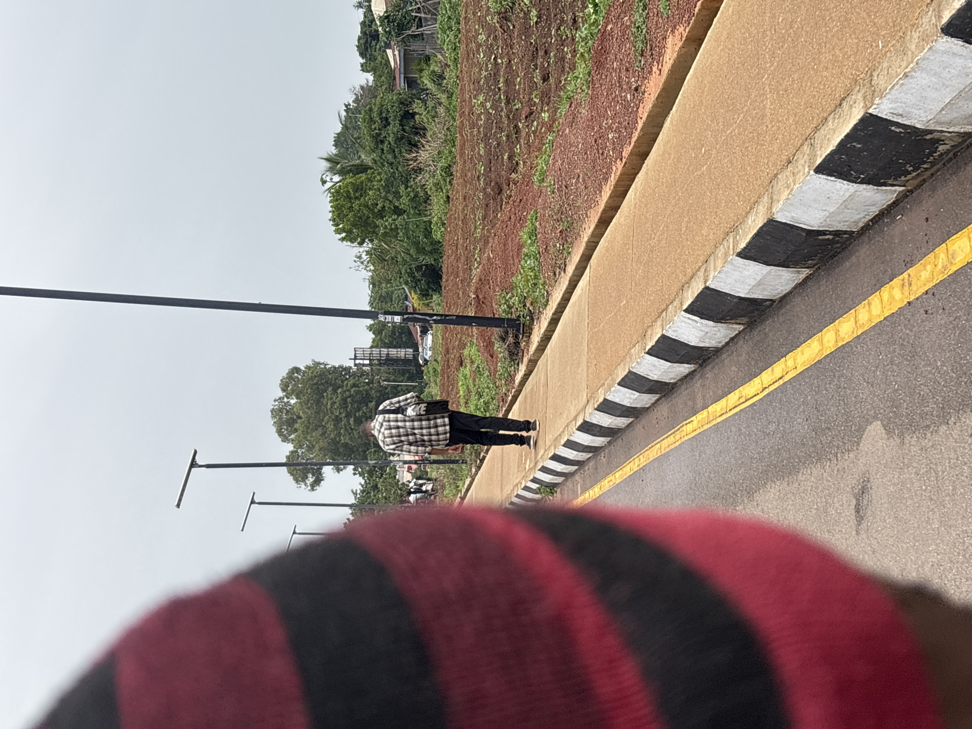 A person walks along a paved sidewalk next to a road in Jos, Nigeria. The person is wearing a black and white checkered shirt and black pants. They are carrying a black bag with white lettering that appears to say "NIKE". The sidewalk is bordered by a curb painted with black and white stripes. The road has a yellow line marking its edge. Beyond the sidewalk is a field with reddish-brown soil and sparse green vegetation. In the background, trees and some buildings are visible, suggesting a semi-urban or rural environment. Several streetlights with horizontal arms are also present. The sky is overcast, indicating a cloudy day. The image appears to be taken from a low angle, possibly from someone walking or moving alongside the subject. The foreground is dominated by the blurred red and black stripes of clothing, likely belonging to the person taking the photo. There is no clear indication of the time of day other than it is daylight.