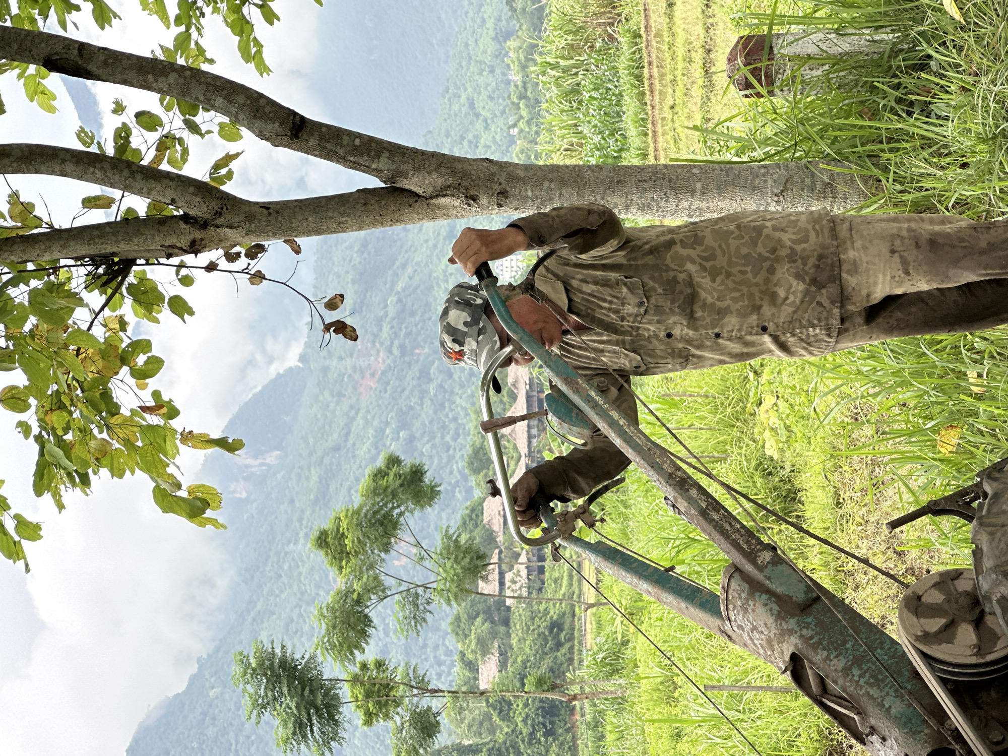 An older man in a camouflage cap with a red star and a camouflage patterned shirt stands in a grassy field, holding onto the handles of a green, weathered tiller. His pants are also stained with dirt. The man appears to be adjusting or operating the tiller, which is positioned in front of him. The background reveals a lush, green landscape with rice paddies, tall grasses, and trees. In the distance, mist-covered mountains rise, characteristic of the scenery in Mai Chau, Vietnam. The sky is overcast with clouds.