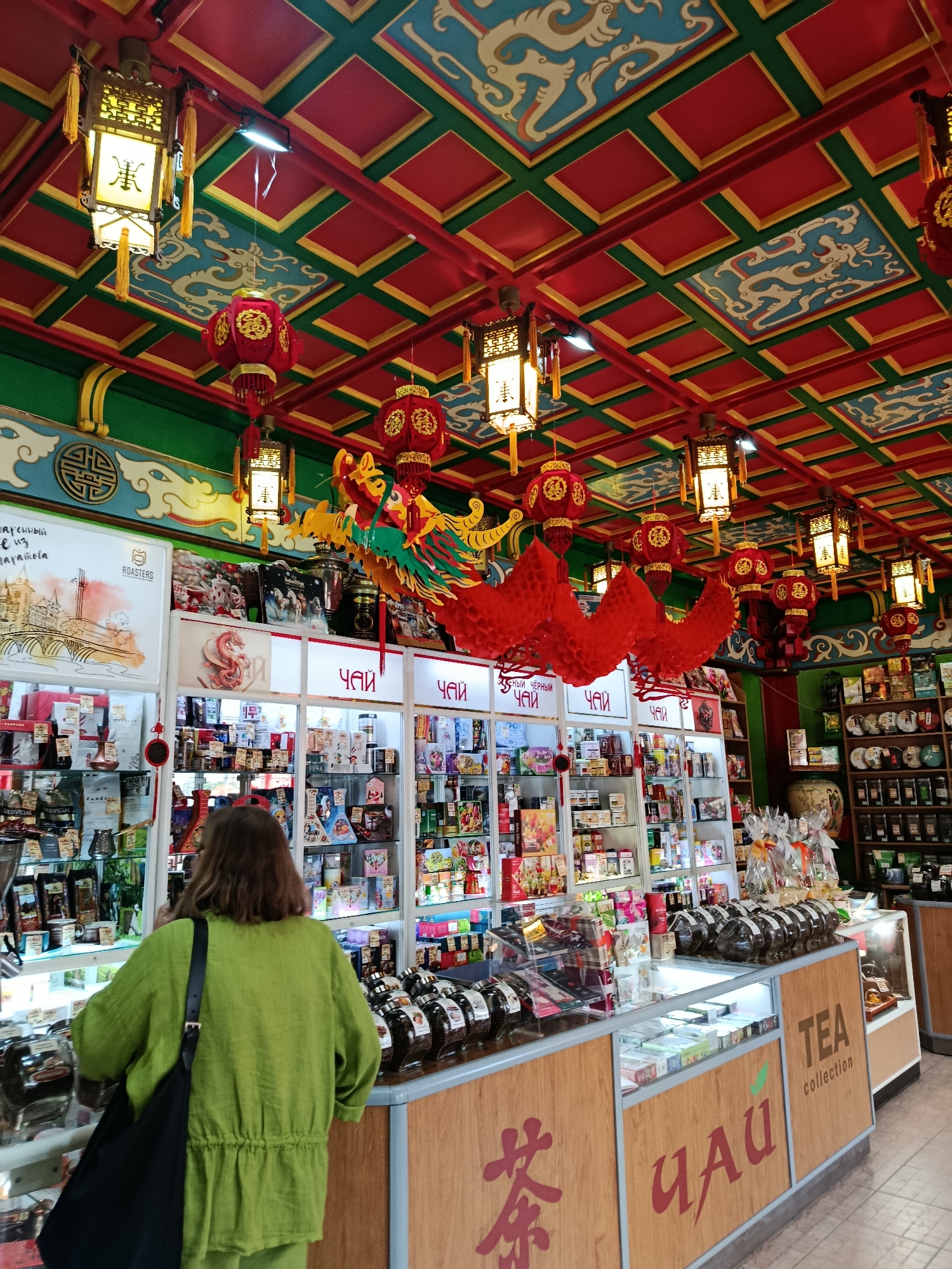 The image depicts the interior of a tea shop, likely located in Saratov, Russia, as indicated by the presence of Russian text on some of the merchandise and signage. The ceiling is elaborately decorated with a red and green grid pattern, interspersed with blue panels featuring white dragon motifs. Numerous Chinese-style lanterns, in red and yellow, hang from the ceiling, as well as decorative red paper decorations and a colorful dragon effigy.
In the foreground, a person wearing a bright green linen outfit and carrying a black shoulder bag is facing away from the camera, looking at displays of tea products. The shop is well-stocked with shelves and display cases filled with various teas, packaged goods, and other related items. The displays feature a mix of products in boxes, jars, and bags. Some signage in Cyrillic indicates "ЧАЙ" (tea) and "TEA collection." Wooden display counters and shelves are visible throughout the shop. The overall impression is a vibrant and culturally themed retail environment.