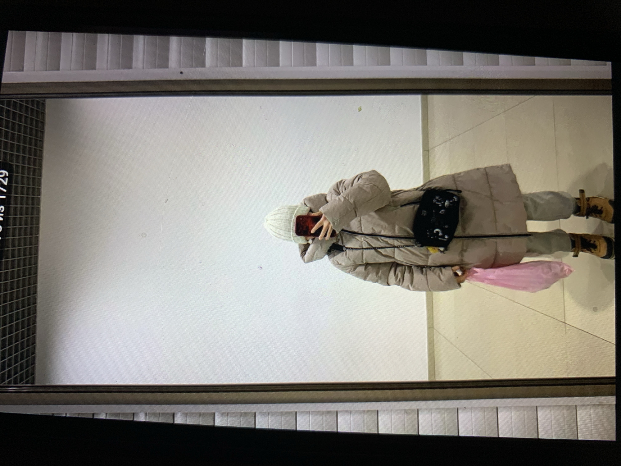 In Lymanka, Ukraine, a person stands in front of a floor-to-ceiling mirror in what appears to be an indoor public space, possibly a mall or store. The person is wearing a light-colored, quilted puffer coat, light-colored pants, brown lace-up boots, and a white knitted beanie. They are holding a pink plastic bag in their left hand and a red-cased smartphone in their right hand, which is being used to take a selfie. A black crossbody bag with a floral pattern is slung across their body. The background of the mirror reflection shows a plain white wall and a tiled floor. The surrounding environment includes a dark, textured frame on the left and right sides of the mirror, and a grid-like ceiling.