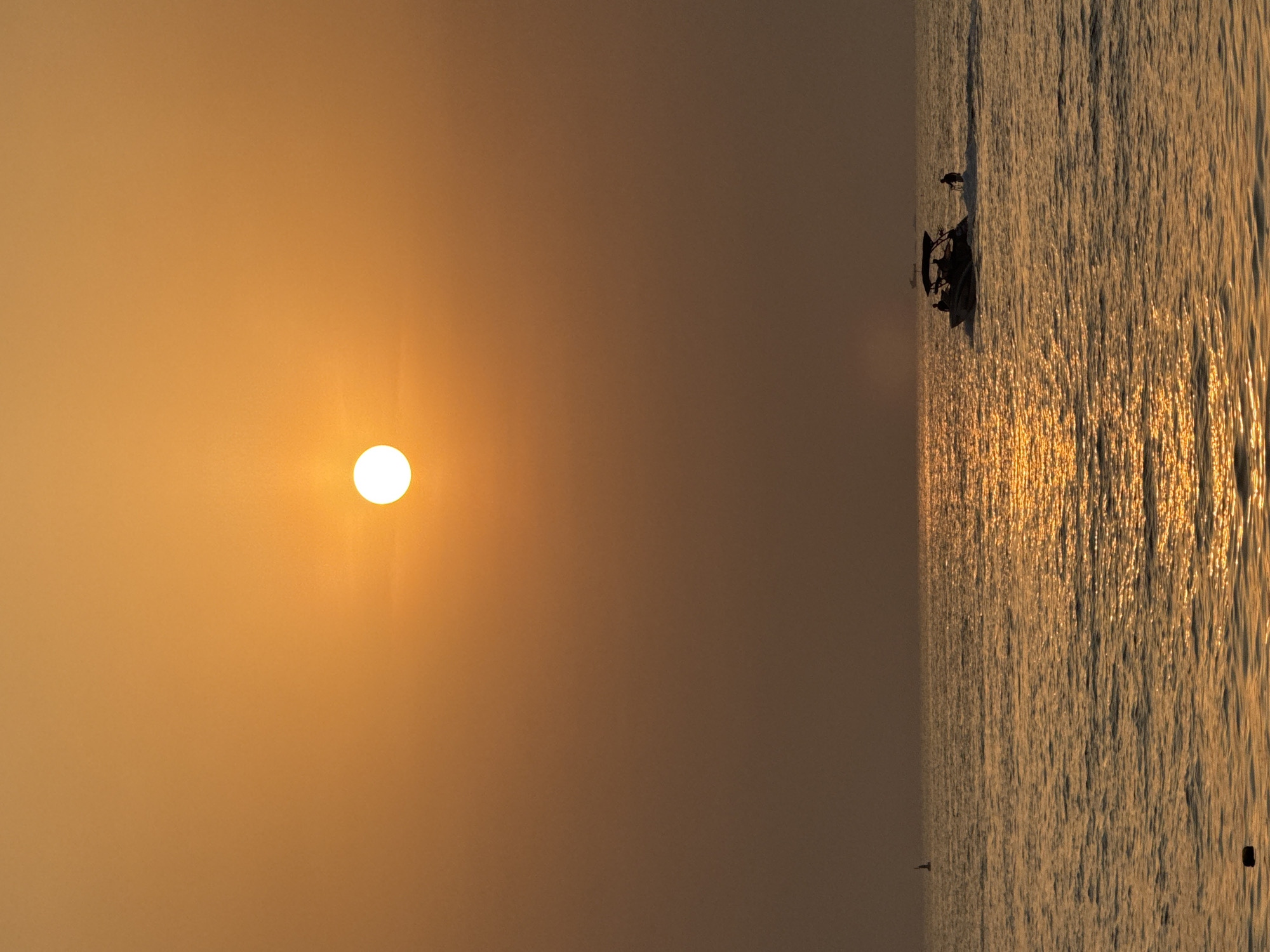 The image shows a serene sunset over the Arabian Gulf in Dubai, United Arab Emirates. The sun, a bright orange orb, is positioned high in the hazy sky, casting a warm glow across the water. The sea is calm, with gentle waves reflecting the golden light. A speedboat, carrying several passengers, cruises across the water, leaving a white wake. In the distance, a lone sailboat is visible on the horizon. The overall atmosphere is peaceful and tranquil, evoking the beauty of a Dubai sunset.