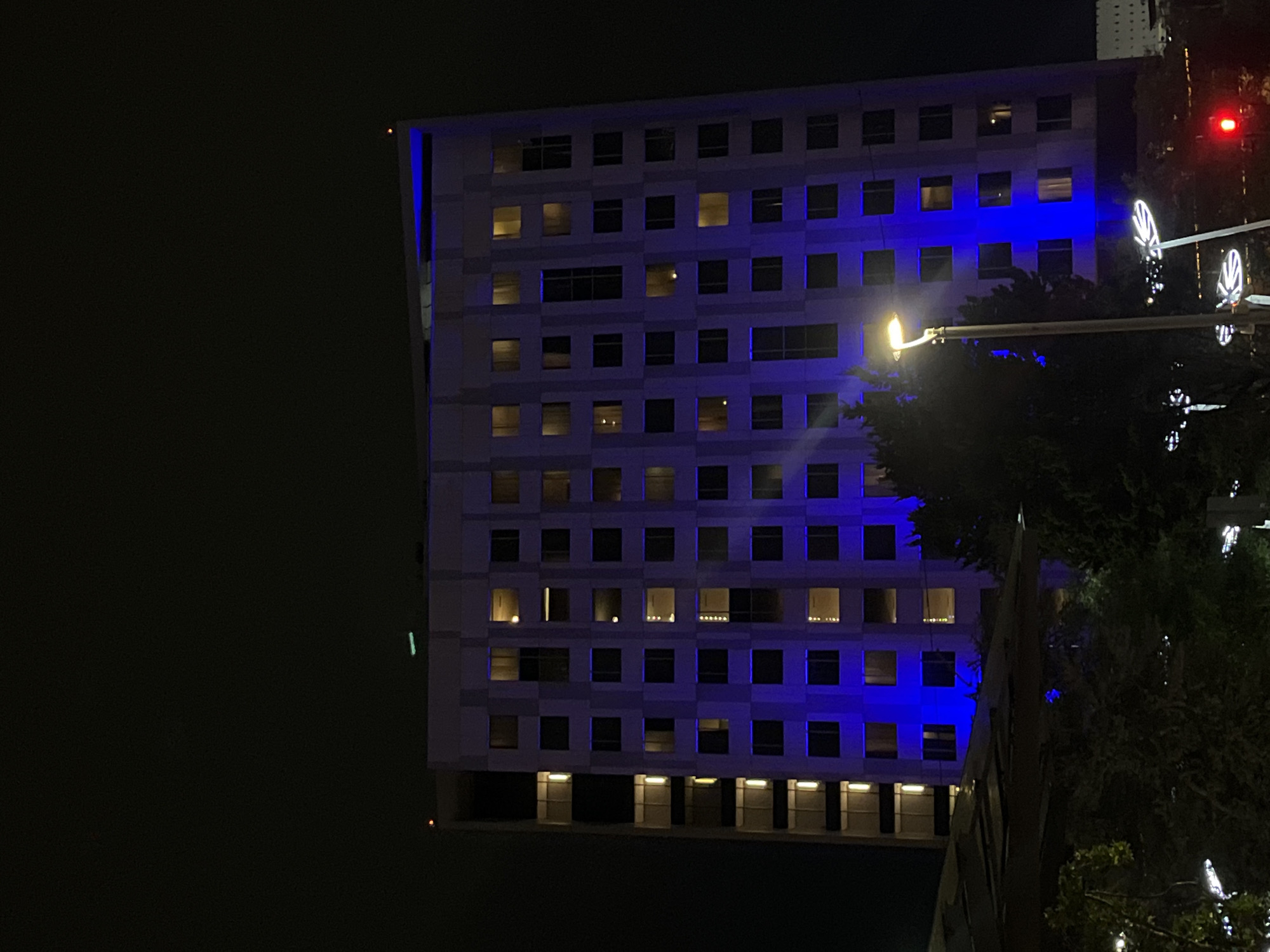 The image shows a building at night. The building is illuminated with blue lights and has several floors with windows. There is a streetlight in front of the building and some trees to the right. A traffic light is also visible. The location is Unknown, Unknown.