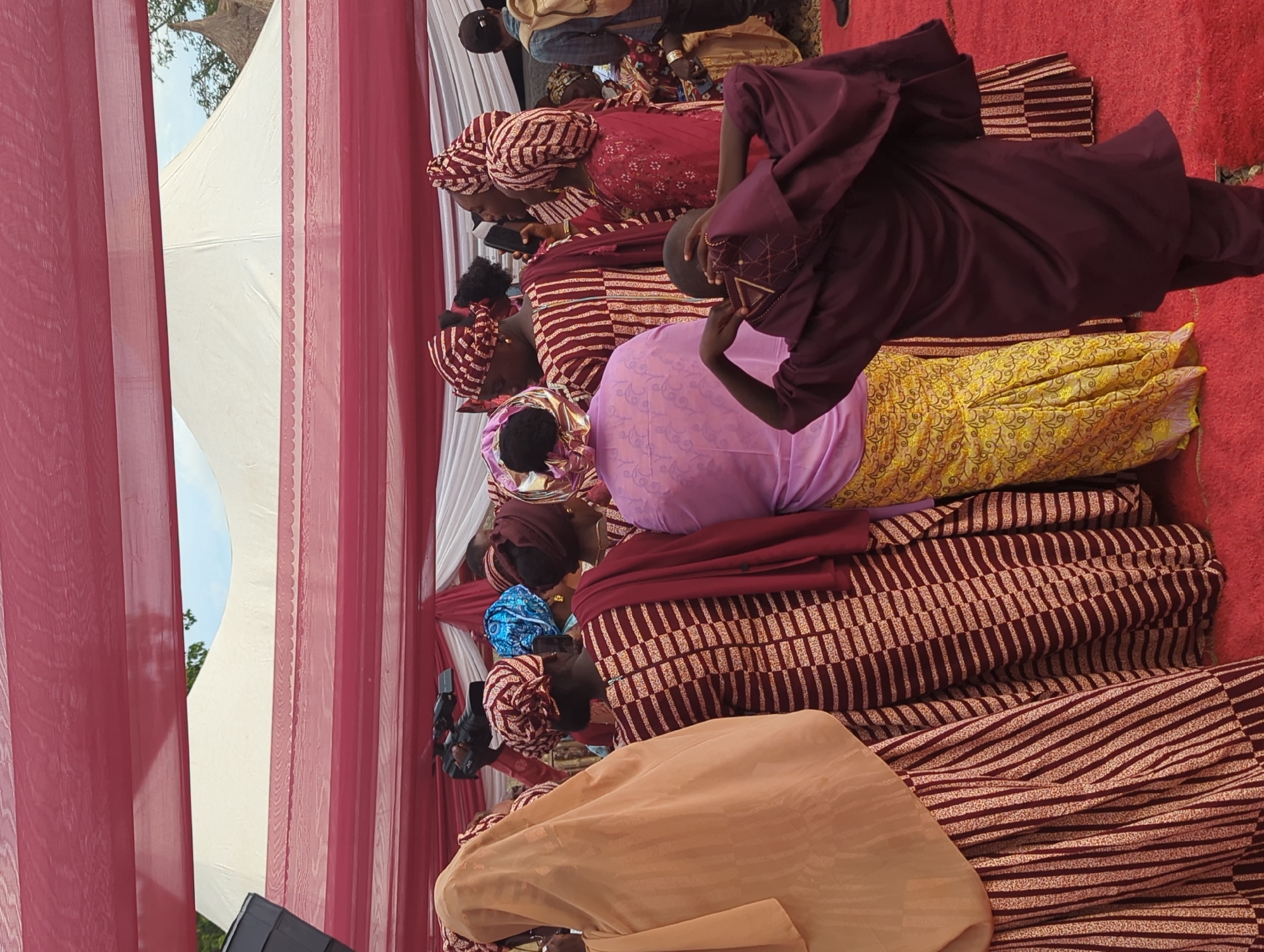 This image depicts an outdoor gathering taking place in Kabala, Nigeria, under a temporary canopy structure. The ground is covered with a red carpet.
The scene is populated by approximately 10-15 visible individuals, primarily women, dressed in various forms of traditional attire. Many women wear patterned dresses and matching head wraps in shades of maroon, red, and light tan, featuring vertical stripes or geometric patterns.
- On the far left, a woman is seen from the back, wearing a light tan head covering and a vertically striped dress.
- In the center-left, a woman is visible from the back, wearing a maroon and tan patterned dress with a dark red shawl draped over her shoulders. Her head is covered with a matching patterned wrap.
- Slightly behind her, a person is operating a black JVC video camera on a monopod or tripod, pointing towards the right.
- In the central foreground, another woman is viewed from the back, wearing a light purple top and a yellow patterned skirt, with a matching purple head wrap.
- To the right, several other women in similar attire are present, some engaged in conversation. One woman in a red dress is holding a black smartphone to her ear. Another woman wears a distinctive bright blue head wrap.
- In the immediate foreground on the right, a young individual, possibly a child or adolescent, is observed from behind, wearing a dark maroon tunic with light embroidery near the neckline and matching pants. Their hands are raised to their head, seemingly adjusting headwear.
- In the far right background, a person with a black baseball cap and a light-colored backpack is partially visible.
The overhead canopy consists of alternating red and white fabric sections. In the background, a large white marquee-style tent is visible, with parts of a blue sky and green foliage, including the top of a traditional straw-roofed structure, peeking out from behind it.