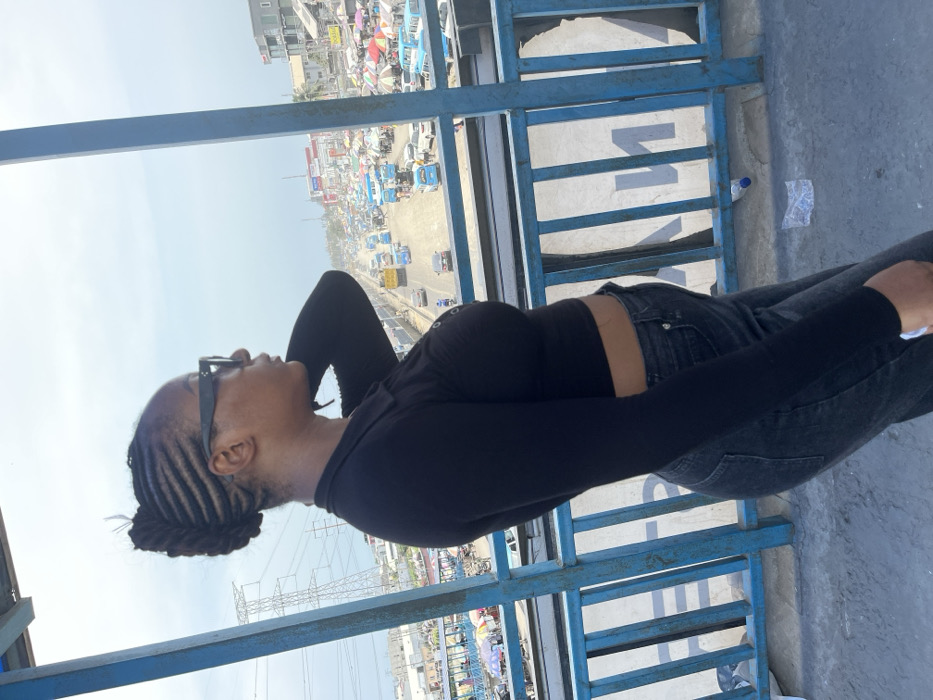 The image depicts a female individual standing on a pedestrian bridge overlooking a busy urban street in Lekki, Nigeria. The subject, visible from mid-thigh up and facing right in profile, has dark skin, hair styled in cornrows pulled back into an updo, and wears dark rectangular sunglasses. She is dressed in a black long-sleeved, collared, cropped top with front button details and dark-wash jeans. Her left arm is raised with the hand positioned behind her head/neck.
The bridge features blue metal railings with vertical bars. On the concrete bridge floor near the bottom right, a clear plastic water bottle and a crumpled plastic bag are present.
Below the bridge, a multi-lane expressway is visible with moderate to heavy vehicle traffic, including passenger cars, a prominent yellow truck, and numerous light blue public transport buses. To the right of the expressway, a dense market area is established, characterized by a multitude of white and brightly colored umbrellas and canopies covering numerous stalls, with multi-story buildings beyond. To the left of the expressway, scattered buildings and a tall electricity transmission tower (pylon) are visible. The sky is light grey-blue, indicative of an overcast or hazy atmospheric condition. The individual appears to be observing the activity below the bridge.