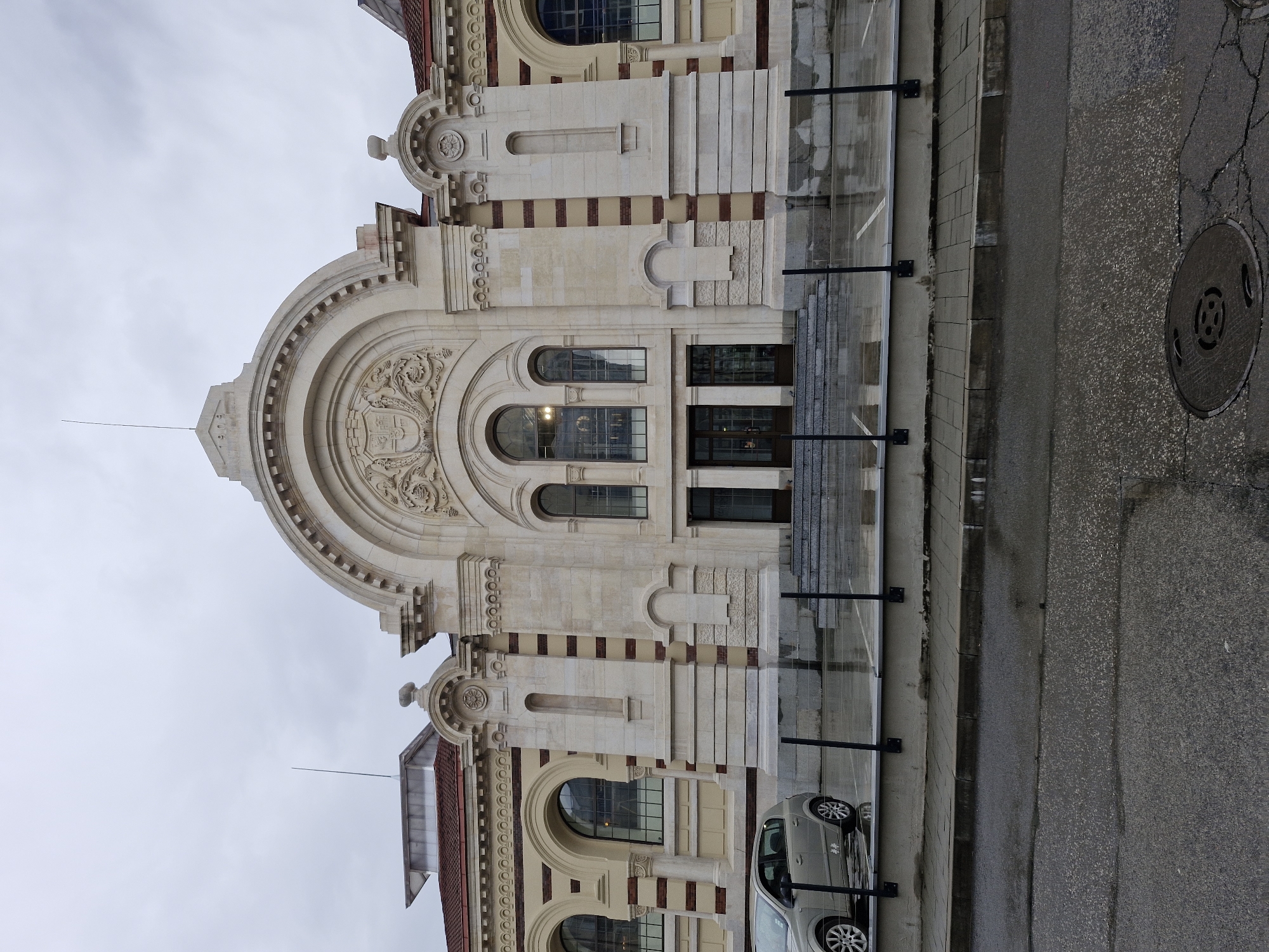 The image captures the facade of the Central Sofia Market Hall in Sofia, Bulgaria. The building displays elaborate architectural detailing with arched windows, decorative stone carvings, and a prominent curved pediment topped with a spire. A glass barrier runs along the edge of the building, separating it from the street. A silver car is parked adjacent to the building. The asphalt street shows signs of wear. The sky is overcast.