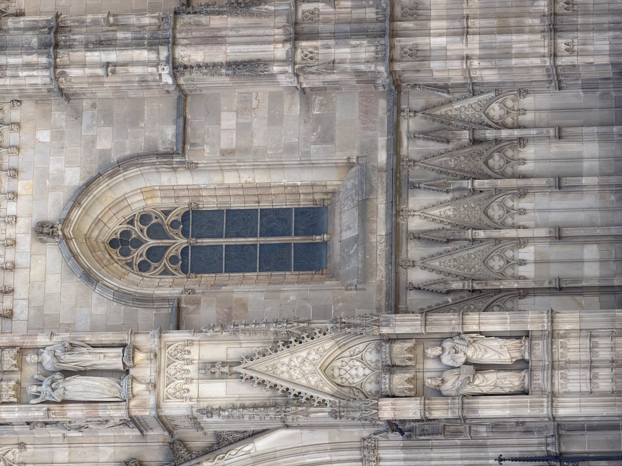 This is a medium shot of the facade of the Barcelona Cathedral in Barcelona, Spain, showing Gothic architecture. The facade is made of light-colored stone blocks. In the upper left, there are two sculpted figures in niches. The figure on the left appears to be an angel with wings. The figure on the right is a bearded man holding an object. Below these figures is a large Gothic window with intricate tracery and dark blue glass. To the right of the window is a vertical architectural element with fluted columns. Below the window, there is a row of decorative stone pinnacles and archways. In the lower half of the image, there are more elaborate carved stone elements, forming a series of pointed arches and Gothic detailing. The lighting suggests it is daytime with soft shadows, indicating overcast or diffused sunlight.