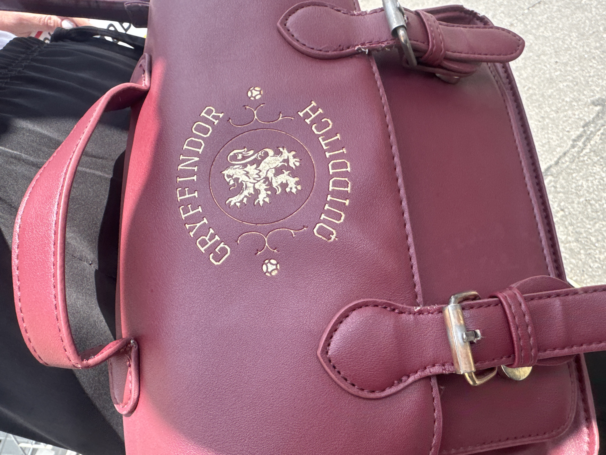 The image shows a close-up of a burgundy-colored satchel with gold lettering and a crest. The satchel appears to be made of faux leather and features buckles and straps for closure. The gold emblem on the satchel is circular and contains an image of a lion, with text around it. The visible text on the satchel reads "GRYFFINDOR" and "QUIDDITCH".
A person's hand with white nail polish is visible in the upper left corner of the image, holding the satchel. The person is wearing dark-colored pants. The background is slightly blurred but appears to be an outdoor setting with a textured surface, possibly a sidewalk or paved area. Sunlight casts shadows on the satchel, indicating it is daytime. The overall scene suggests a casual outing or travel.
The context provided indicates the location is Kremenchuk, Ukraine, and it is implied to be a sunny day, possibly during warmer months. No specific emotions are evident beyond what might be inferred from the general context of holding and displaying a personal item.