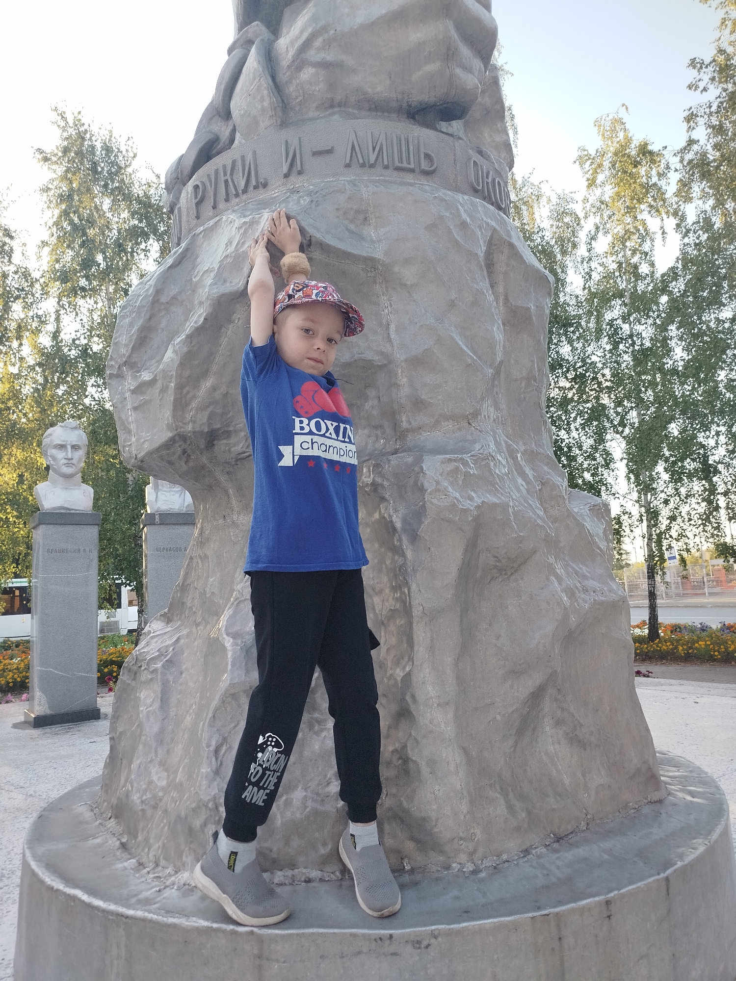 In Yalutorovsk, Russia, a young boy stands in front of a large concrete monument with carved text. He is wearing a blue t-shirt with "BOXING Champion" printed on it, black pants with white lettering that reads "BACK TO THE GAME", and gray sneakers. The boy is gripping the monument with both hands, with his arms extended upwards. Behind him, the monument appears to be part of a larger sculpture or installation. Two busts on pedestals are visible to the left of the boy, and trees with green foliage line the background. The scene is outdoors, with a clear sky and sunlight illuminating the area.