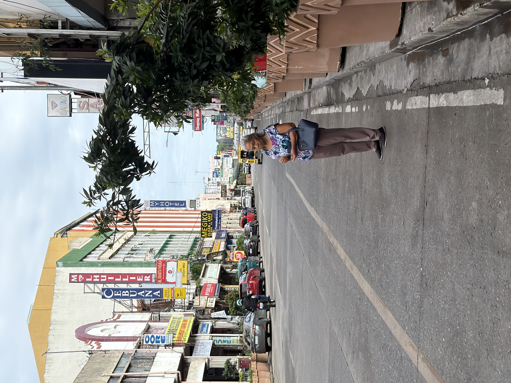 The image captures a street scene in Davao City, Philippines. An elderly woman stands on the sidewalk next to a planter. The street is filled with parked cars and motorcycles. Various signs line the buildings, advertising businesses such as pawn shops, money transfer services, and hotels. Notable signs include "CEBUANA," "M LHUILLIER," "CORE," "MY HOTEL," and "MEGIKO." The street appears to be a commercial area with a mix of businesses.