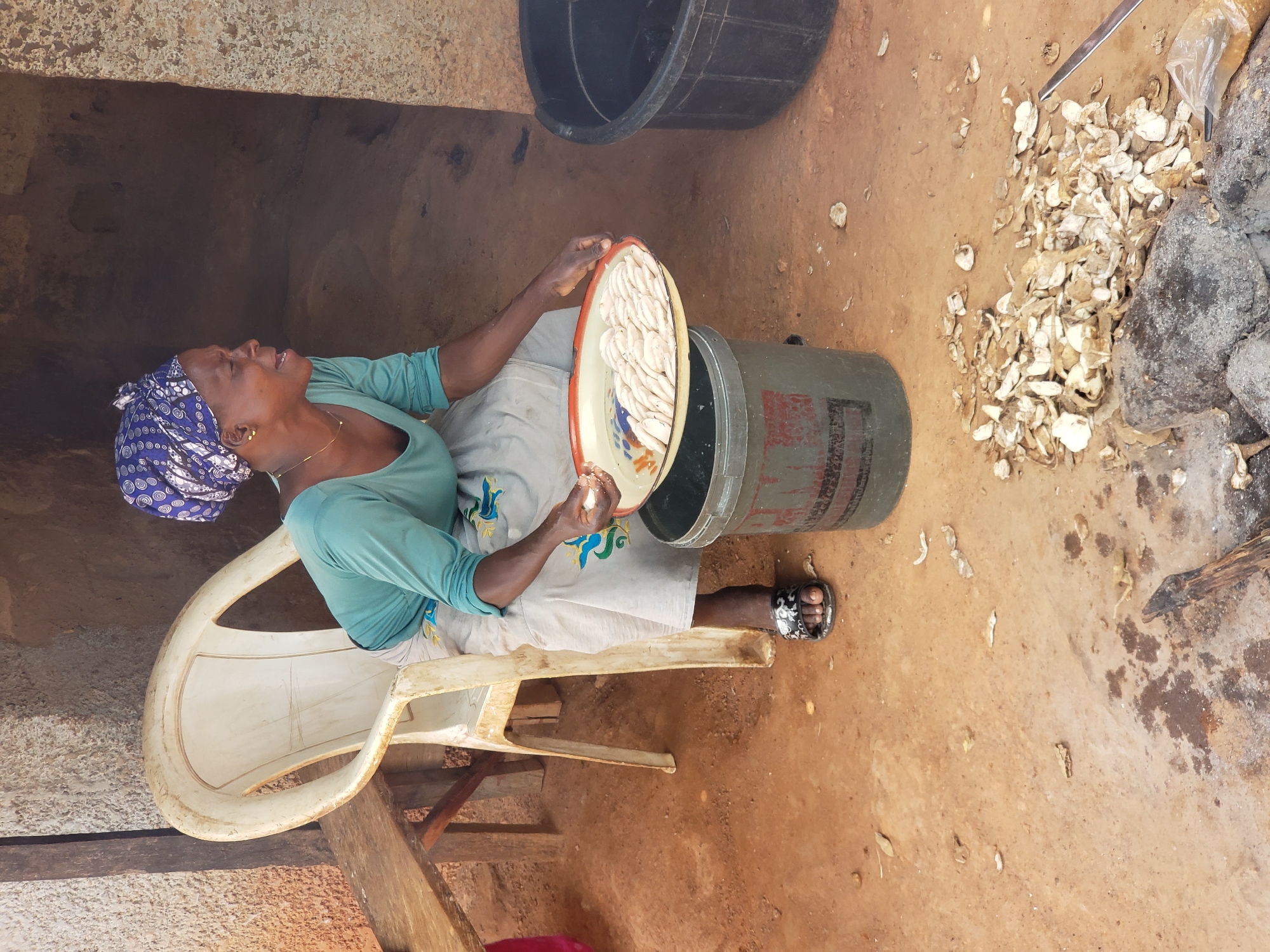 In Narayi, Nigeria, a woman sits on a white plastic chair, holding a shallow, round metal bowl filled with white, cylindrical food items. She is wearing a teal blouse, a patterned headscarf, and a light-colored skirt. Her right hand is in the bowl, and she appears to be in the process of preparing food. Beside her is a large green bucket, into which she is about to place or has just taken the bowl. To the right of the bucket, a large black plastic container is visible. The ground is covered in reddish-brown earth, with scattered debris including dried leaves and what appear to be pieces of yam or cassava. In the background, a rough-hewn stone structure and wooden beams suggest an outdoor or semi-enclosed workspace. The scene is illuminated by natural light, casting shadows.