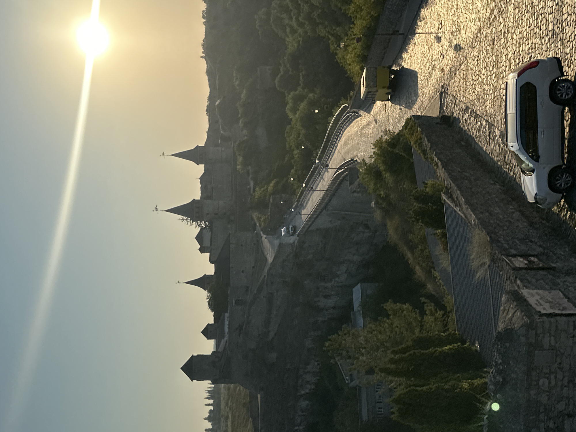 The image shows the Kamianets-Podilskyi Castle in Ukraine at sunset. The castle is a large, stone fortress with multiple towers and battlements, perched on a rocky cliff. A bridge, likely a road, leads to the castle, with a yellow bus and a white SUV driving on it. The sun is low in the sky, casting a warm glow on the scene and creating long shadows. The surrounding landscape is hilly and covered in trees, with a cobblestone road in the foreground. The overall impression is one of historical grandeur and natural beauty.