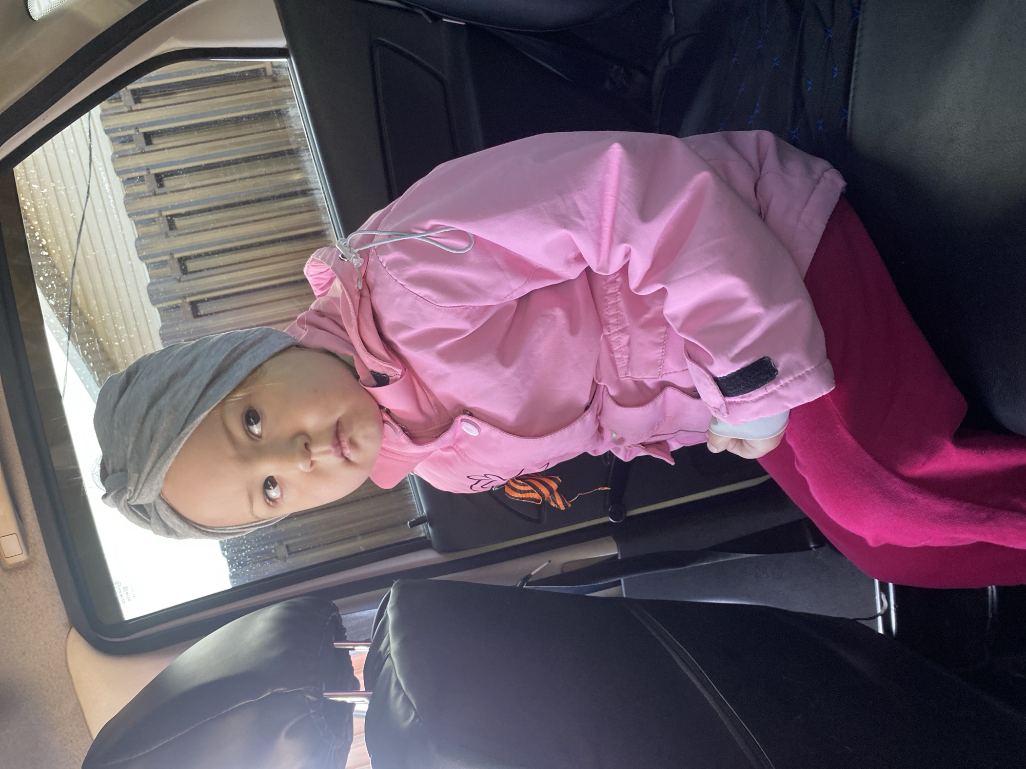 A young child, likely a toddler, is seated in the back of a car in Chaikovsky, Russia. The child wears a light gray headwrap, a bright pink jacket, and dark pink trousers. The pink jacket features a small, embroidered emblem with orange and black stripes, possibly resembling a St. George ribbon, on the upper chest. The child has light-colored eyes and is looking directly at the camera with a neutral, slightly curious expression.
The setting is the interior of a car, indicated by the dark car seats and a headrest visible in the foreground. Through the car window on the left side of the image, streaks of water are visible, suggesting the car is either in a car wash or has recently been exposed to rain. Beyond the window, a light-colored, corrugated metal structure can be seen. The natural lighting indicates it is daytime. Small, white, unreadable text appears on the car windowpane.
