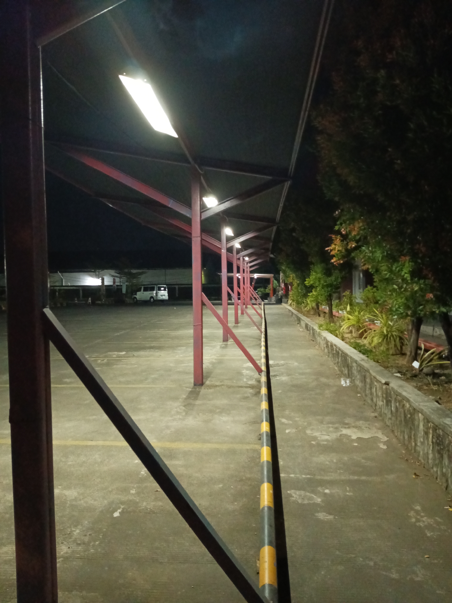 The image depicts a dimly lit outdoor covered parking area at night. The structure is supported by red metal beams and a dark roof, with fluorescent lights illuminating the space. The ground is paved concrete, marked with yellow lines. A concrete wall with some vegetation alongside it runs along the right side of the image. A yellow and black striped barrier pole is visible in the foreground. In the background, a white van is parked in the lot. The scene appears to be located in an urban environment, possibly a parking lot for a commercial or residential complex.