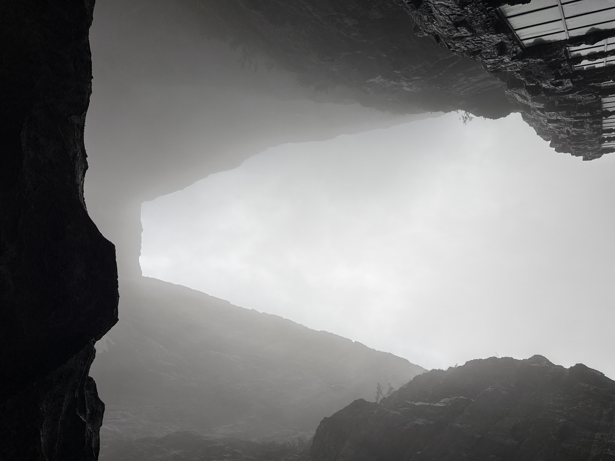 The image depicts a view upwards through a gap in rock formations. Dense fog obscures much of the landscape, but the silhouettes of mountains can be discerned in the middle ground. In the lower right and left corners are dark, rocky cliffs. The upper right corner shows a dark, geometric structure, possibly a building or constructed platform, atop a rock face. The photograph was taken in Yongding District, China.