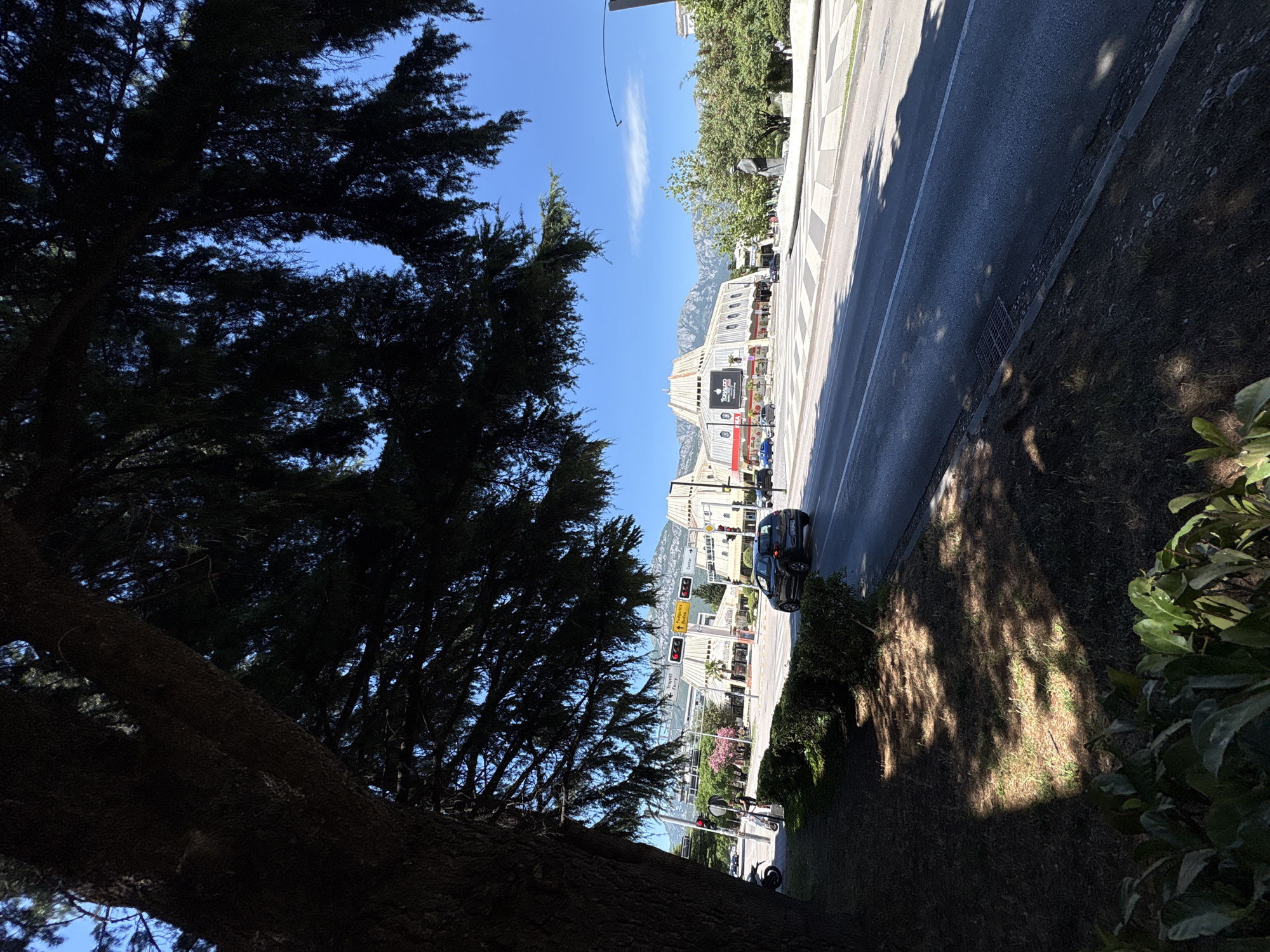 This image captures a street scene in Bar, Montenegro, on a sunny day. In the foreground, a large tree with dark foliage partially obscures the view. The road in front of the tree is a multi-lane asphalt surface, with a white line marking the lanes. To the left of the road, there is a strip of grass and bushes, with dappled sunlight casting shadows. A dark-colored car is driving on the road.
Across the road, a modern building with a distinctive angular roofline dominates the middle ground. The building appears to be a commercial establishment, with large windows and signage that includes the name "Tornado." Several other cars are parked or driving in front of this building. In the background, mountains rise under a clear blue sky with a few wispy clouds. Traffic lights and street signs are visible on the side of the road. The overall impression is of a contemporary urban environment with a backdrop of natural scenery.