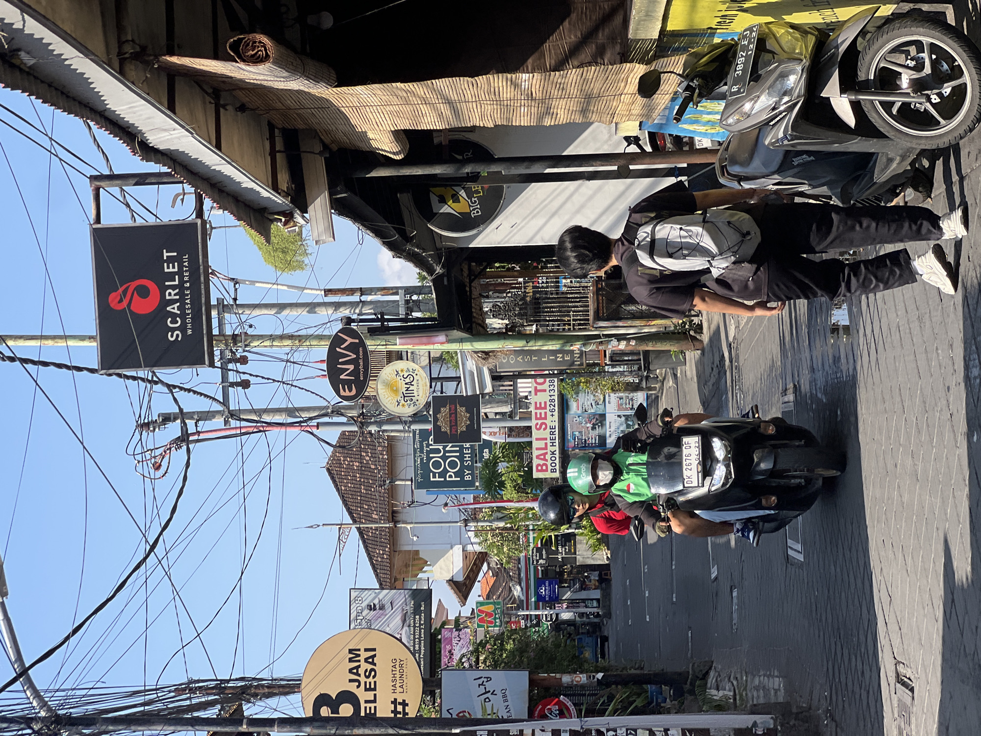The image depicts a street scene in Kuta, Indonesia, under a clear blue sky. A narrow, paved alleyway is lined with buildings and numerous signs, indicating a commercial area. Overhead, a complex network of electrical wires is strung between poles. In the foreground, a person wearing a black t-shirt and black pants with a gray backpack walks away from the camera towards the right side of the image. To the left, a motorcycle with two riders is moving away from the camera. The rider in front wears a green jacket and a white helmet, while the person behind wears a red shirt and a black helmet. The motorcycle has a license plate that reads "DK 2676 QF". Parked on the right side of the alley is another motorcycle. Several signs are visible, including "SCARLET WHOLESALE & RETAIL", "ENVY", "BALI SEE TO...", and "FOUR POINTS BY SHERATON". The buildings exhibit Balinese architectural elements. The overall impression is a bustling street in a popular tourist destination.