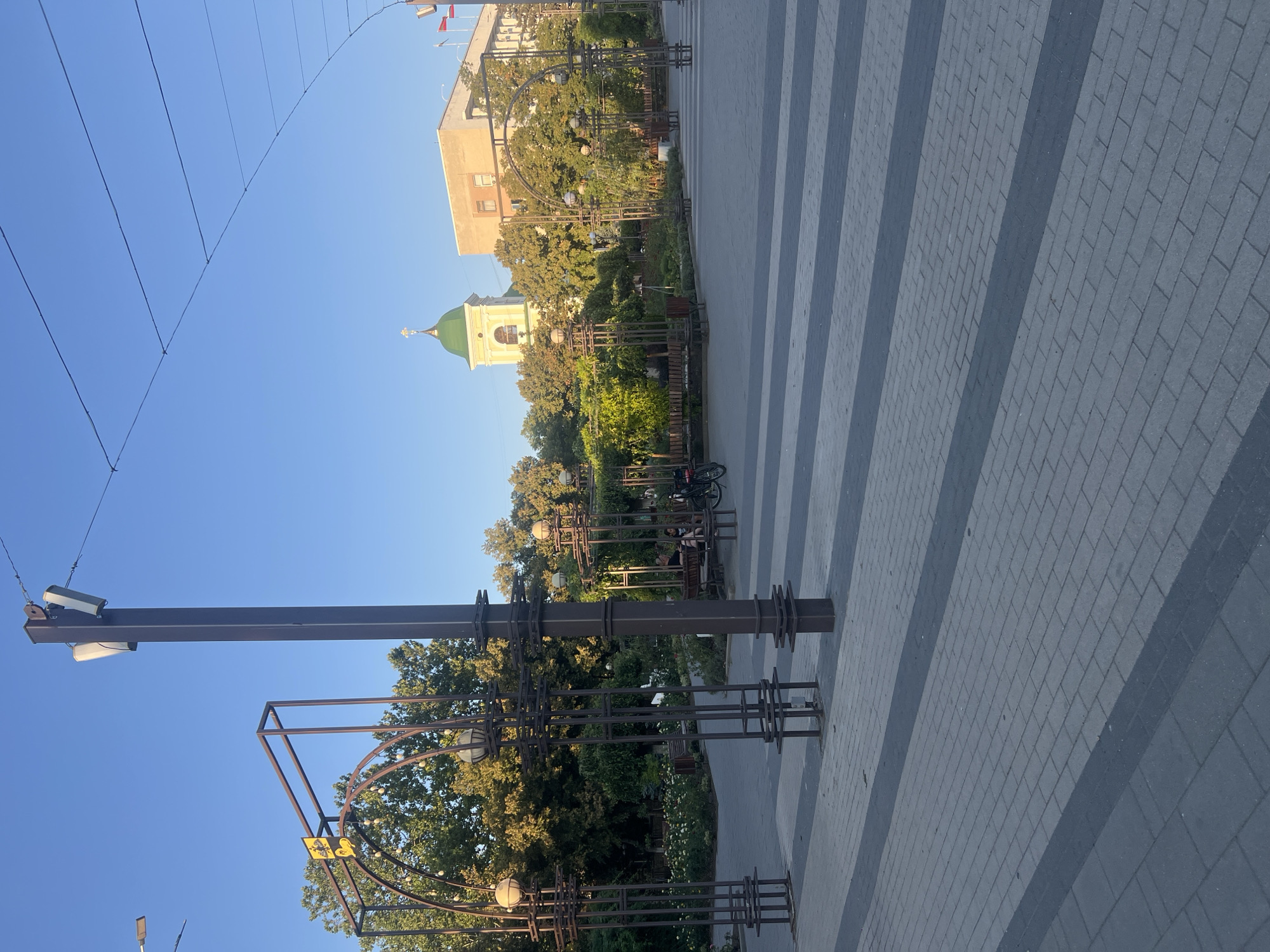 The image depicts a paved public square in the city of Bender, Moldova. In the foreground, the square is paved with grey and dark grey rectangular bricks arranged in parallel stripes. A tall, dark-colored lamppost stands prominently on the right side of the frame, with its top obscured by overhead wires. To the left, a dark metal archway structure is visible, adorned with small spherical lights.
Beyond the square, a park-like area with lush green trees and flower beds is apparent. Several dark metal benches are situated within this area. Two individuals are seated on one of these benches, facing away from the camera. A bicycle is parked nearby.
In the background, a light-colored building with windows stands to the right. To the left of this building and partially obscured by trees, a distinctive yellow bell tower with a green domed roof and a cross on top rises against a clear blue sky. Red and white flags are displayed on poles in front of the building on the right. The overall scene is bathed in the warm light of what appears to be late afternoon or early morning.