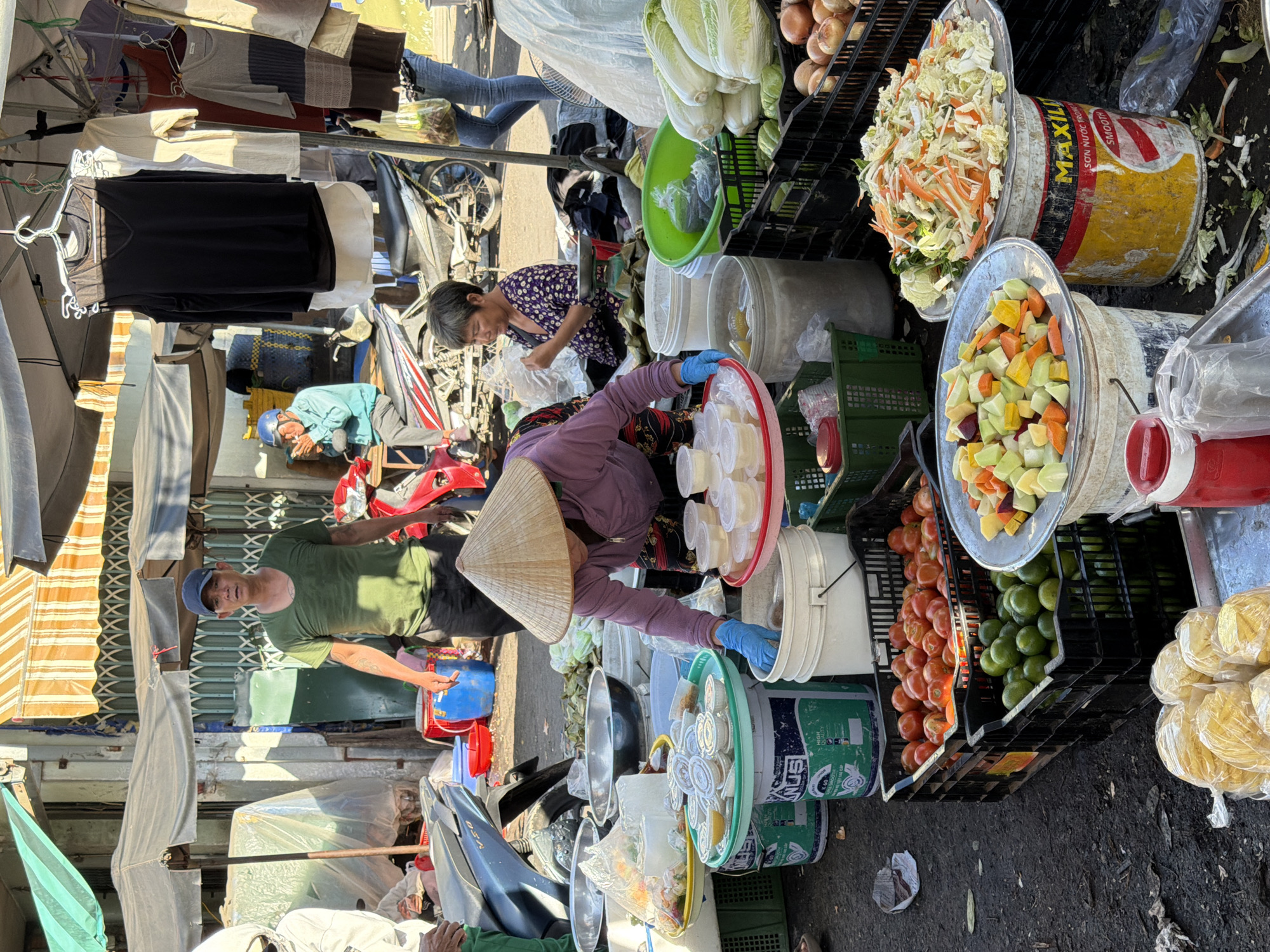 This image captures a bustling street market scene in Nha Trang, Vietnam. A man wearing a green t-shirt and a blue baseball cap stands in the center, appearing to be a vendor. To his right, a woman in a light blue shirt and helmet is seated on a motorcycle, smoking. In the foreground, a woman with a conical hat is arranging small containers of food. Several stalls are visible, displaying a variety of produce including tomatoes, limes, and what appear to be cabbages and onions. Clothing and other goods are also displayed for sale. The overall atmosphere suggests a busy commercial area with daily life unfolding.