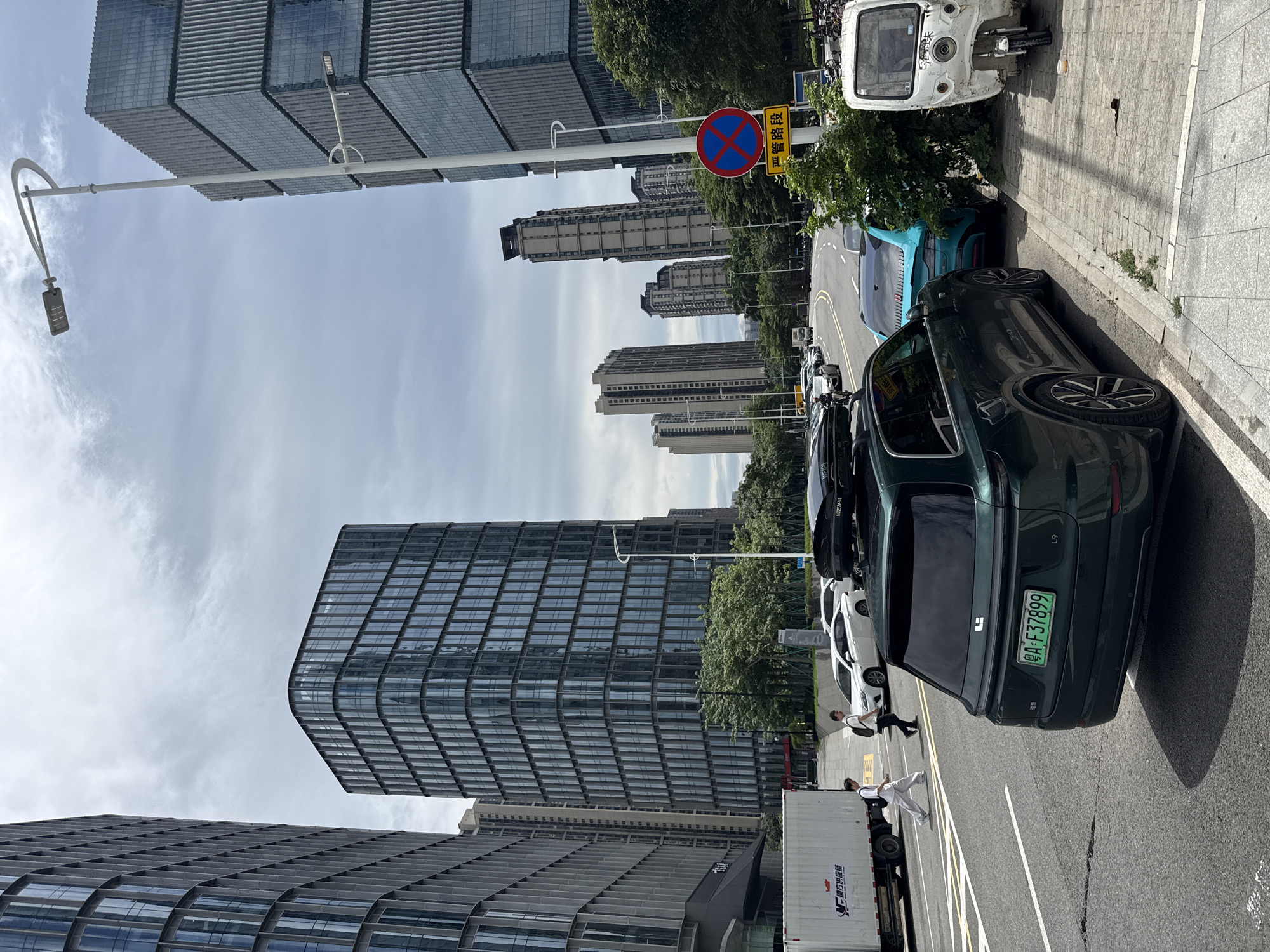 In the Haizhu District of China, the image depicts a street scene with contemporary architecture. On the left, a large, white box truck with Asian characters on its side is parked. Two individuals, one wearing a white shirt and pants and the other a black shirt and pants, are walking across the street, with the person in white appearing to be looking towards the left. A dark green SUV is parked in the foreground, facing away from the camera, with a license plate that reads "A•F37899". The SUV has a roof box. To the right of the SUV, a turquoise car is parked, followed by a white three-wheeled vehicle that resembles a tuk-tuk. Tall, modern glass-fronted buildings dominate the background, with a light-colored sky overhead. A street lamp with two lights is visible on the right side of the frame. A circular red and blue sign with a diagonal cross indicates a prohibition. The road has white lane markings. Green trees are present along the street and sidewalks.