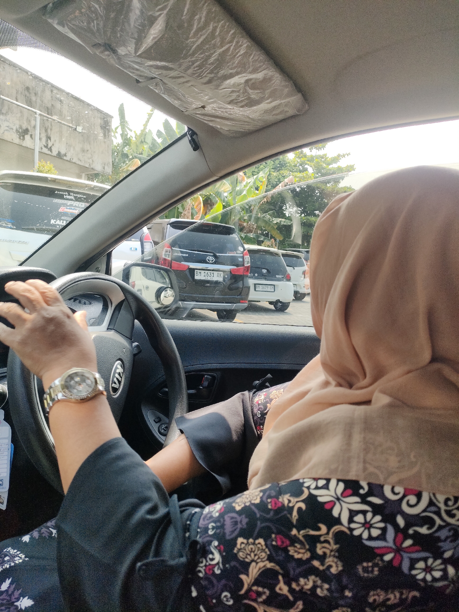 The image, captured from inside a car, shows a person's hands on the steering wheel. The driver is wearing a watch on their left wrist and a black garment with a floral pattern. A woman wearing a light brown hijab is visible in the passenger seat. Outside the car, other vehicles are seen in traffic, including a black SUV with the license plate "BM 1631 AK", and a white car. The setting appears to be a street in Pekanbaru, Indonesia, with a building on the left and trees visible in the background. The sunlight suggests it is daytime.